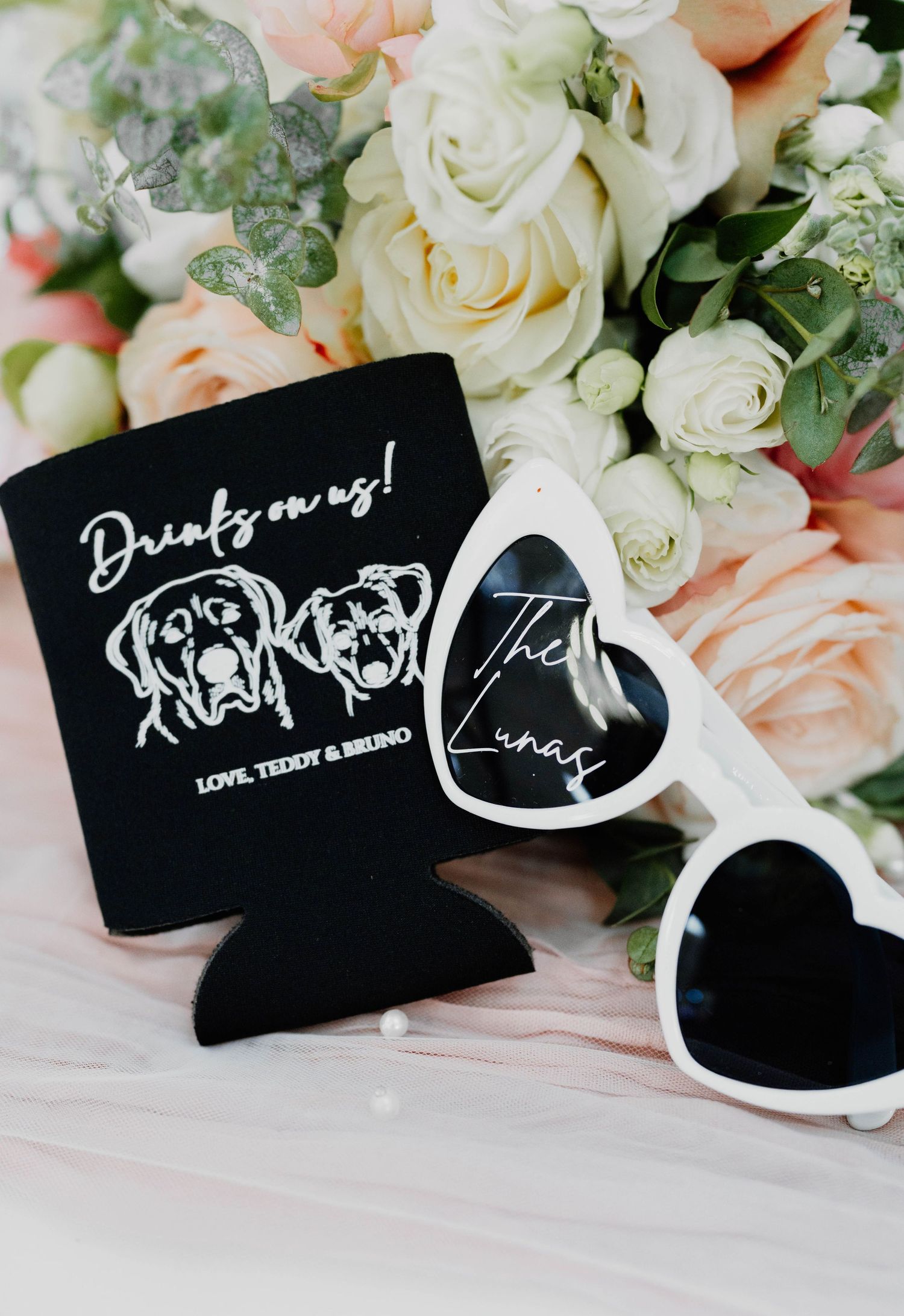 Black wedding koozies with white handwritten text displayed among white roses.