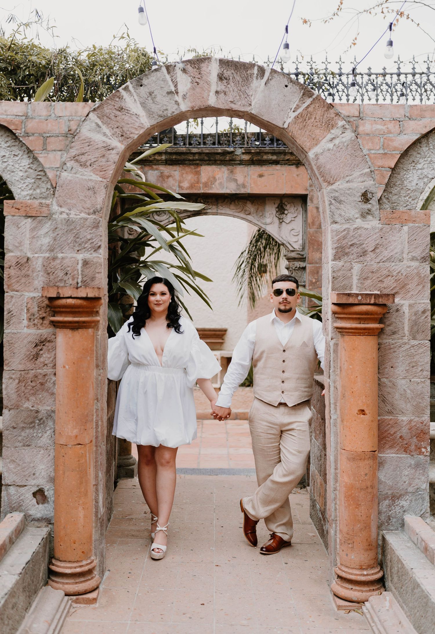 Couple walks and poses beneath stone archways in Mediterranean-style architecture.