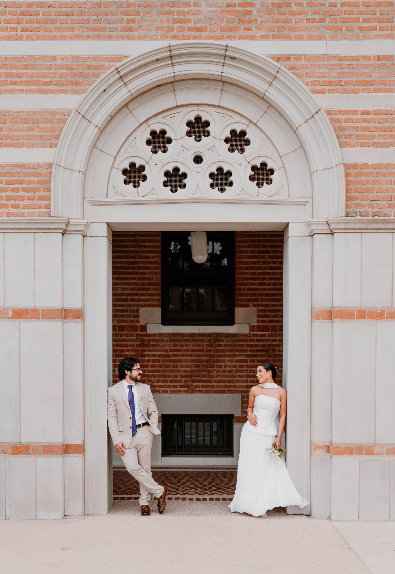 A couple stands in an archway entrance with ornate floral details and brick architecture.