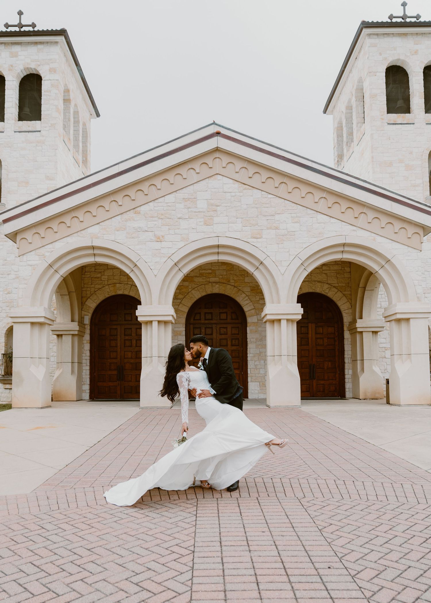 Wedding couple embracing in front of a white church with arched entrances.