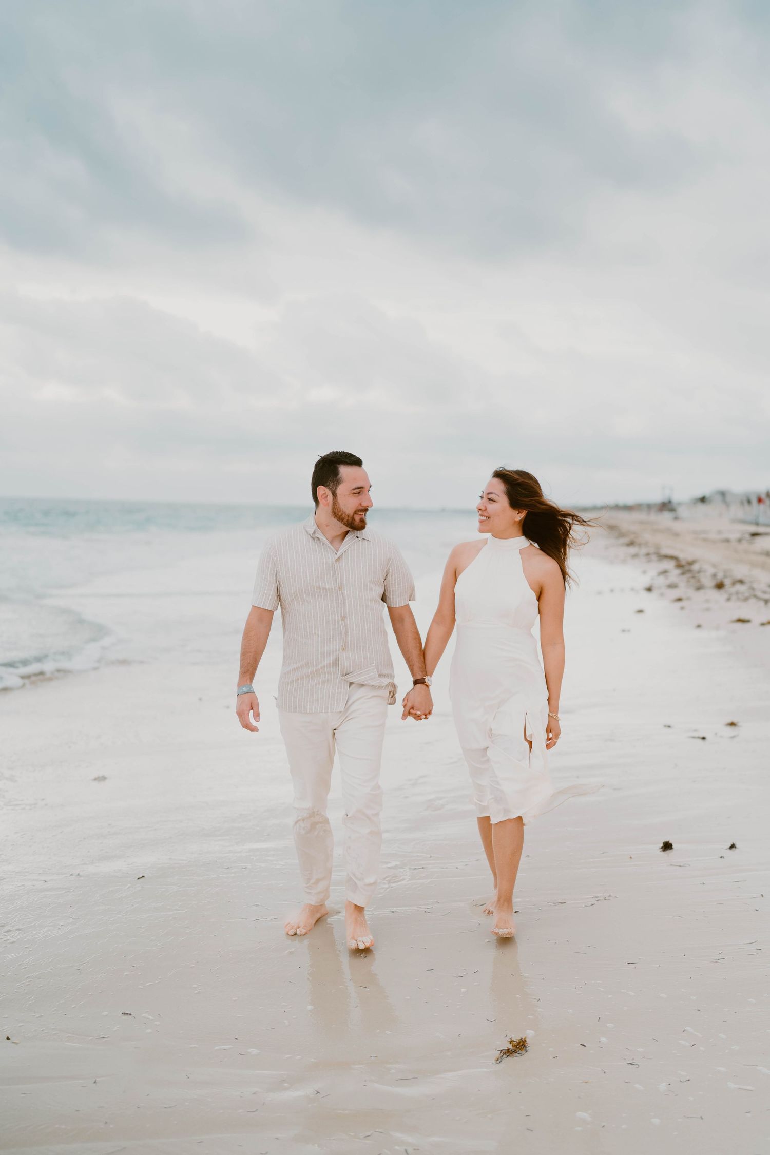 Couple walking barefoot along a misty beach shore in light colored clothing.
