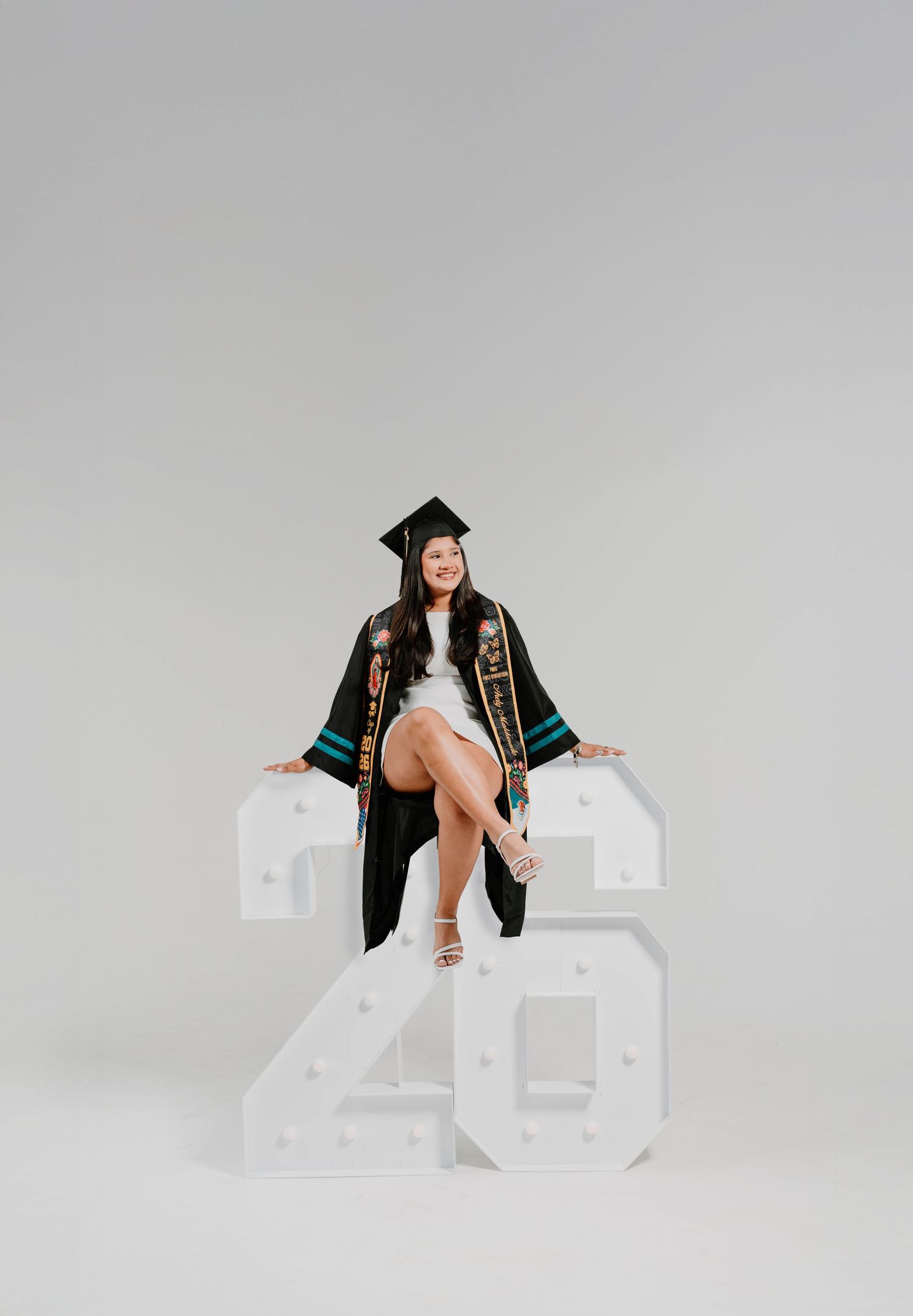Graduate in cap and gown sits on illuminated marquee numbers 25 against a gray studio background.