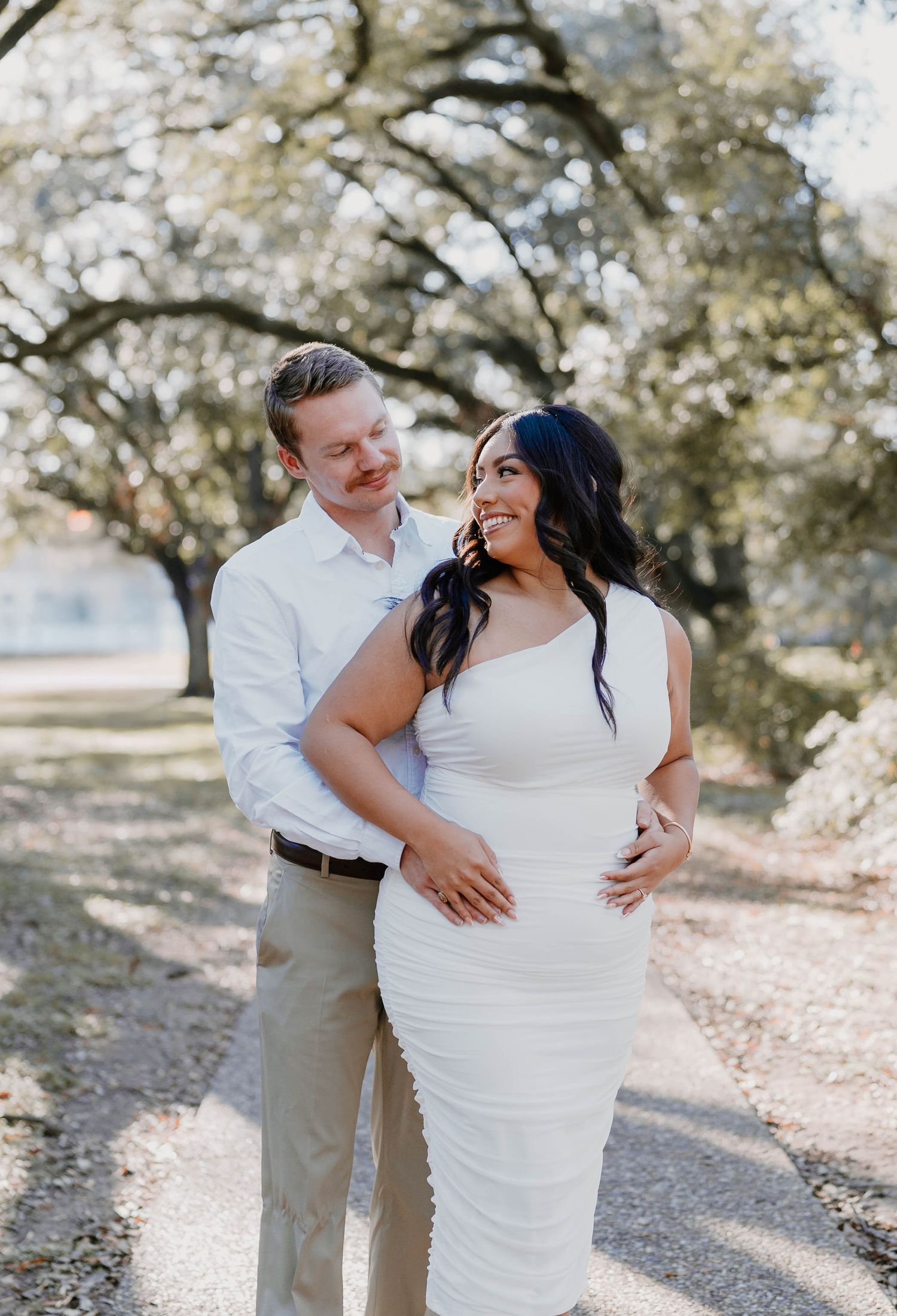 A romantic engagement photo of a couple embracing outdoors under blooming trees on a sunny day.