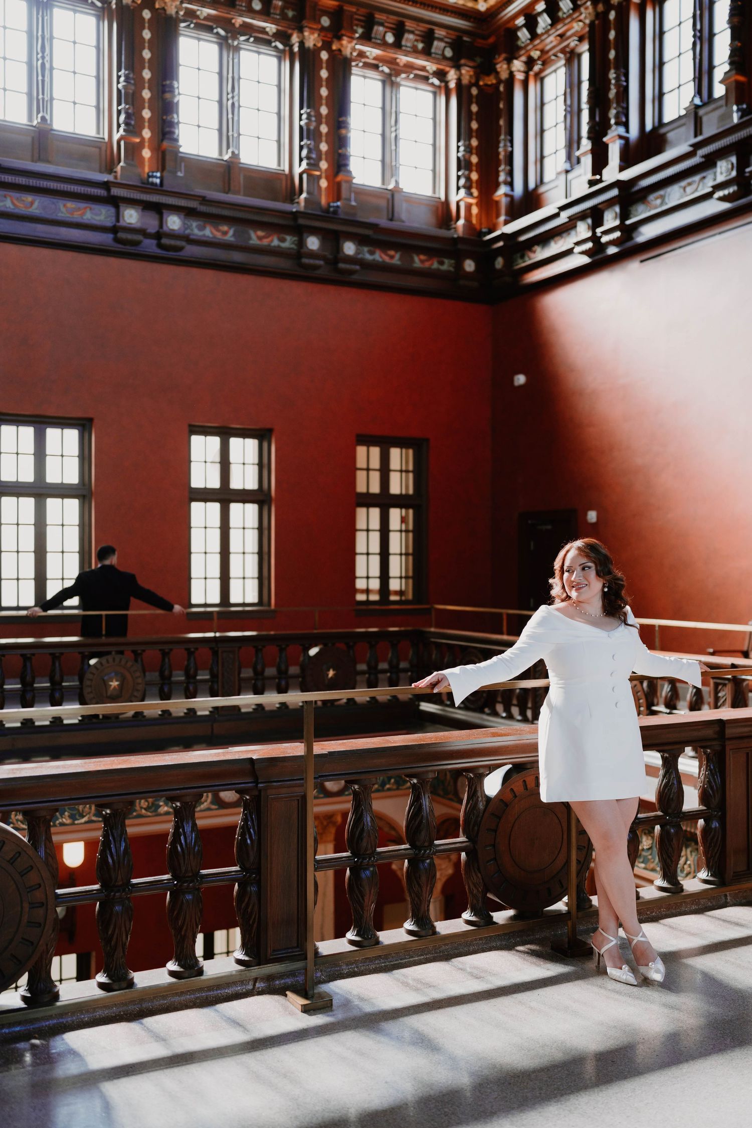 A person in a white dress poses elegantly against a wooden railing in a historic building interior.