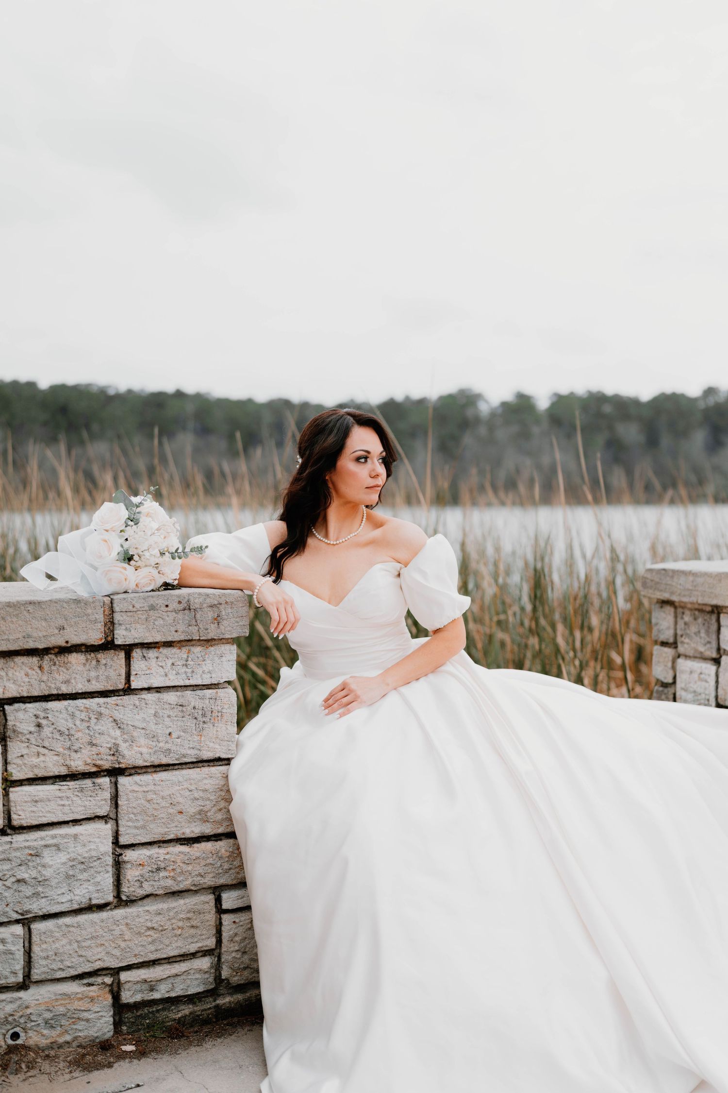 Bride in flowing white dress posing against stone wall near coastal grasses.