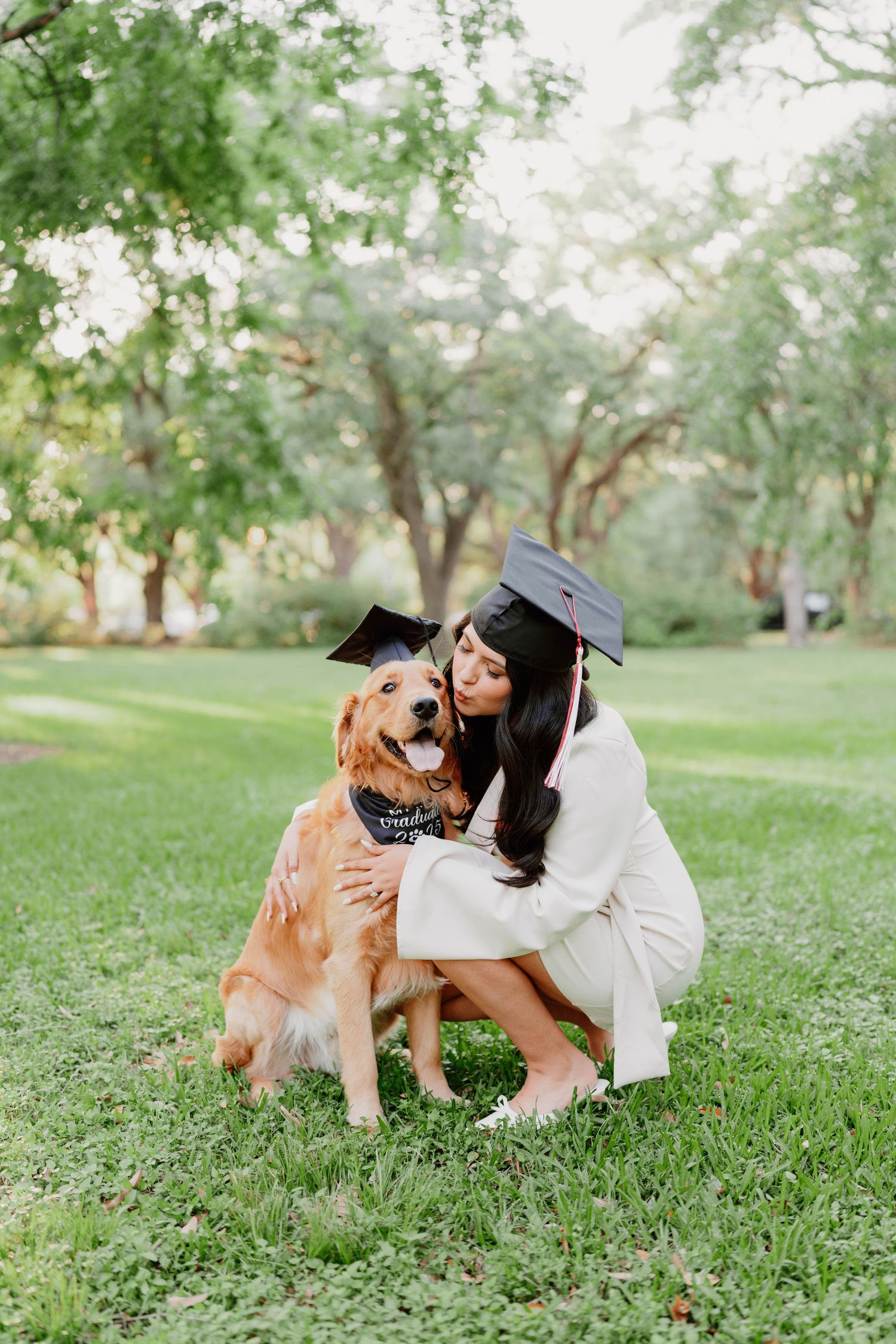 Graduate in white gown cuddles with friendly golden retriever dog in a peaceful park setting.
