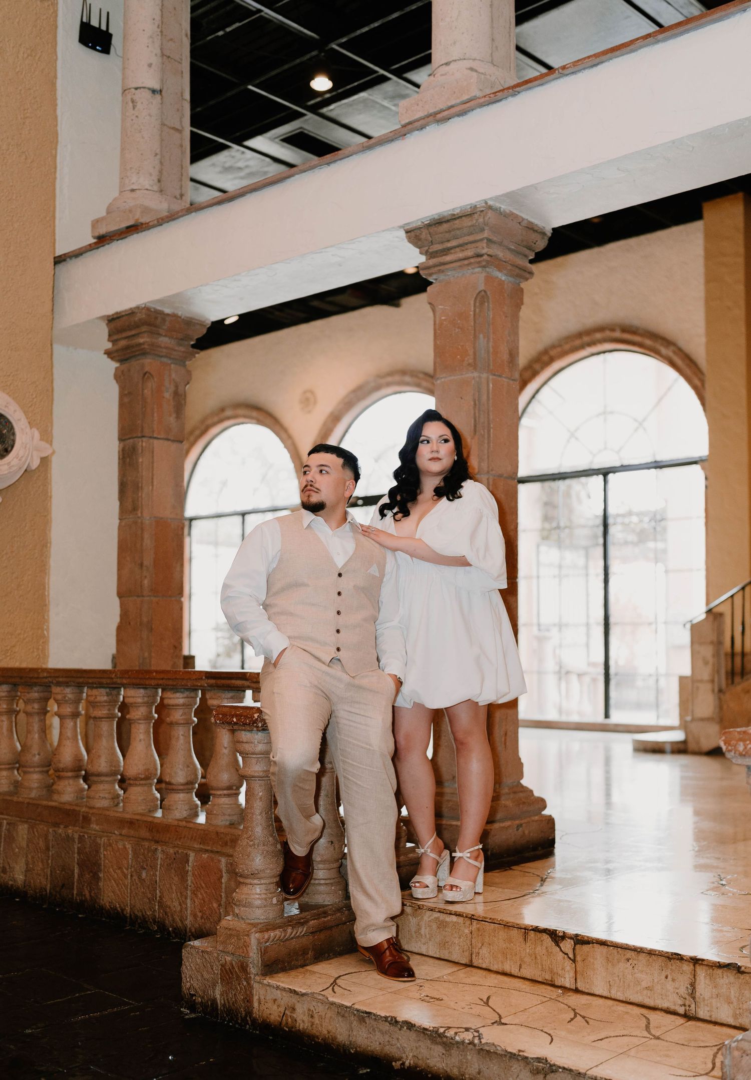 Couple poses in elegant indoor setting with arched windows and stone columns.