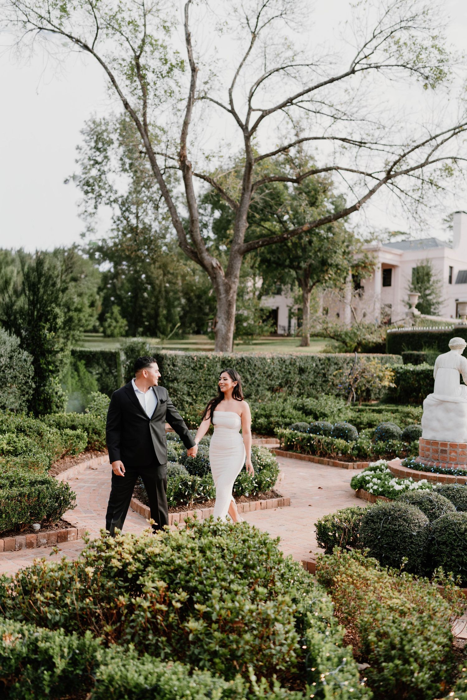 Elegant pair strolls through lush garden with brick pathways and greenery.
