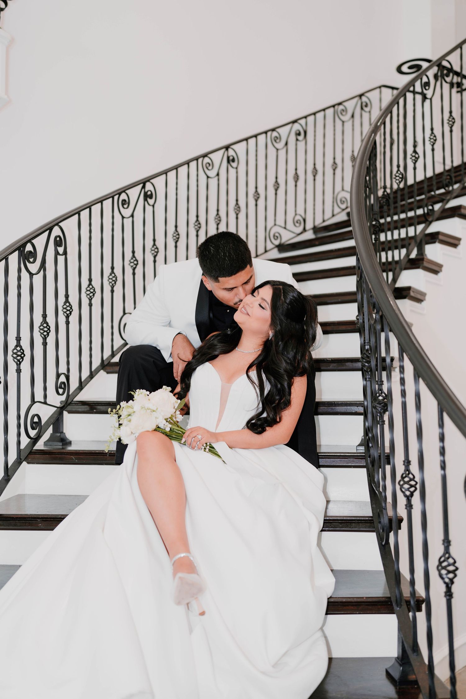 Romantic wedding couple embracing on grand staircase with decorative iron railings.