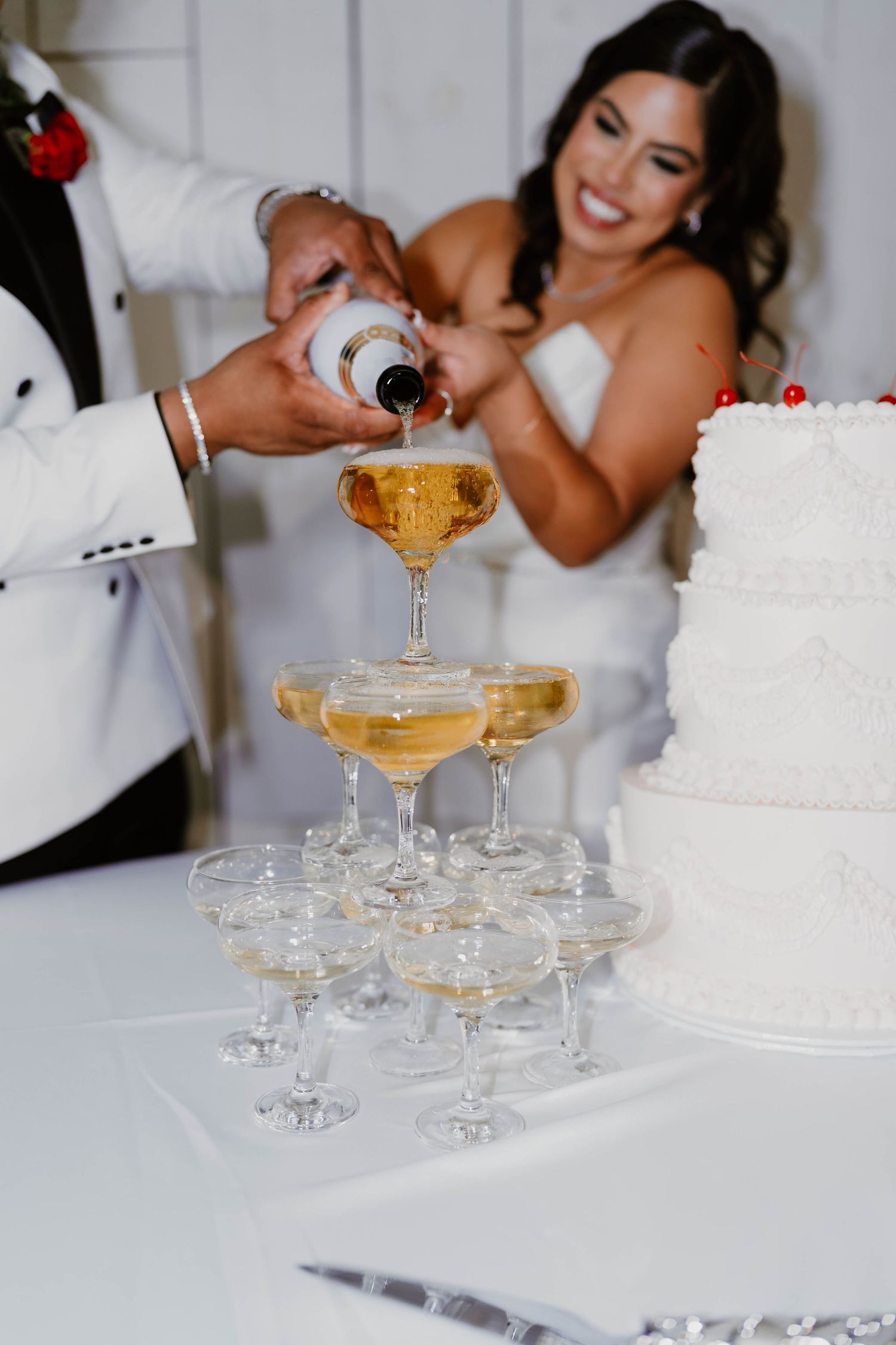 Champagne tower being poured at wedding reception with white cake in background.