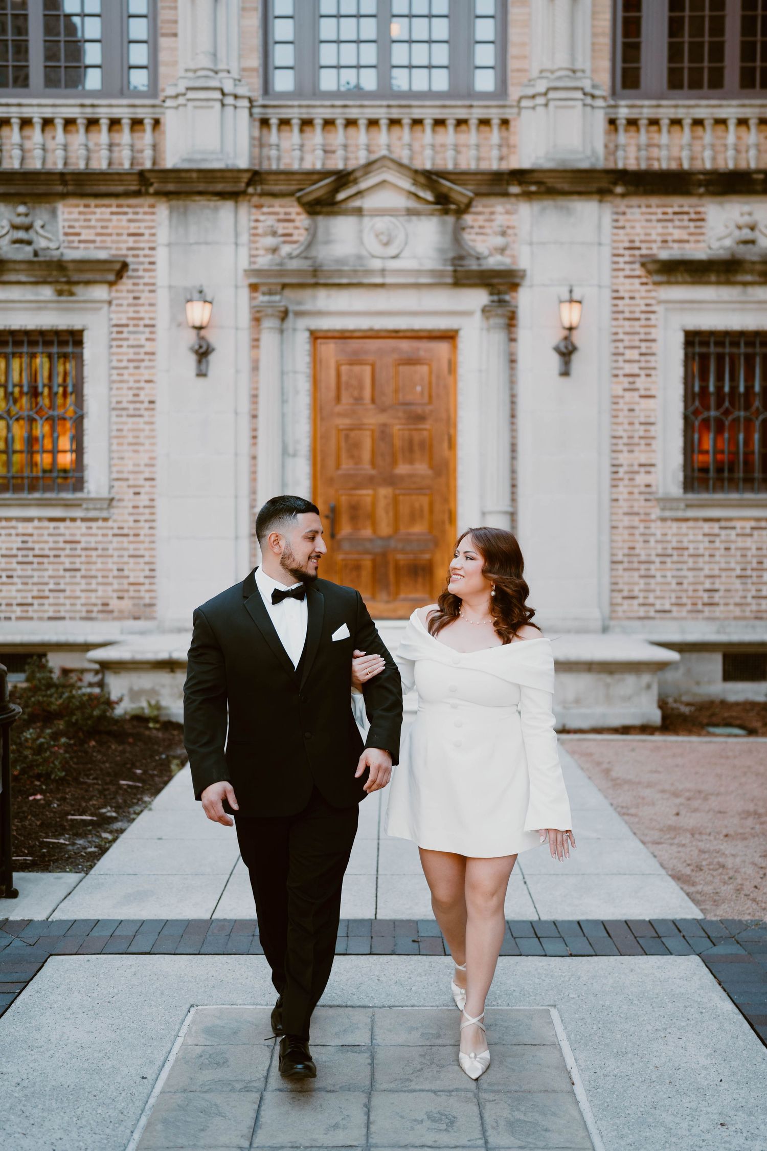 A couple walks together in formal attire outside an elegant brick building with classical details.
