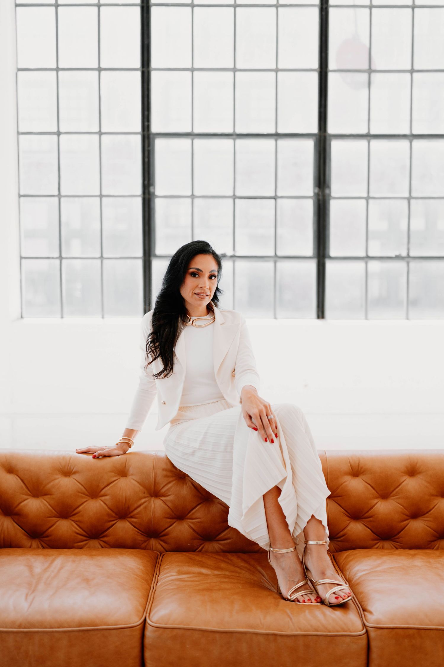 Woman in white dress lounges on brown leather couch in bright studio setting.