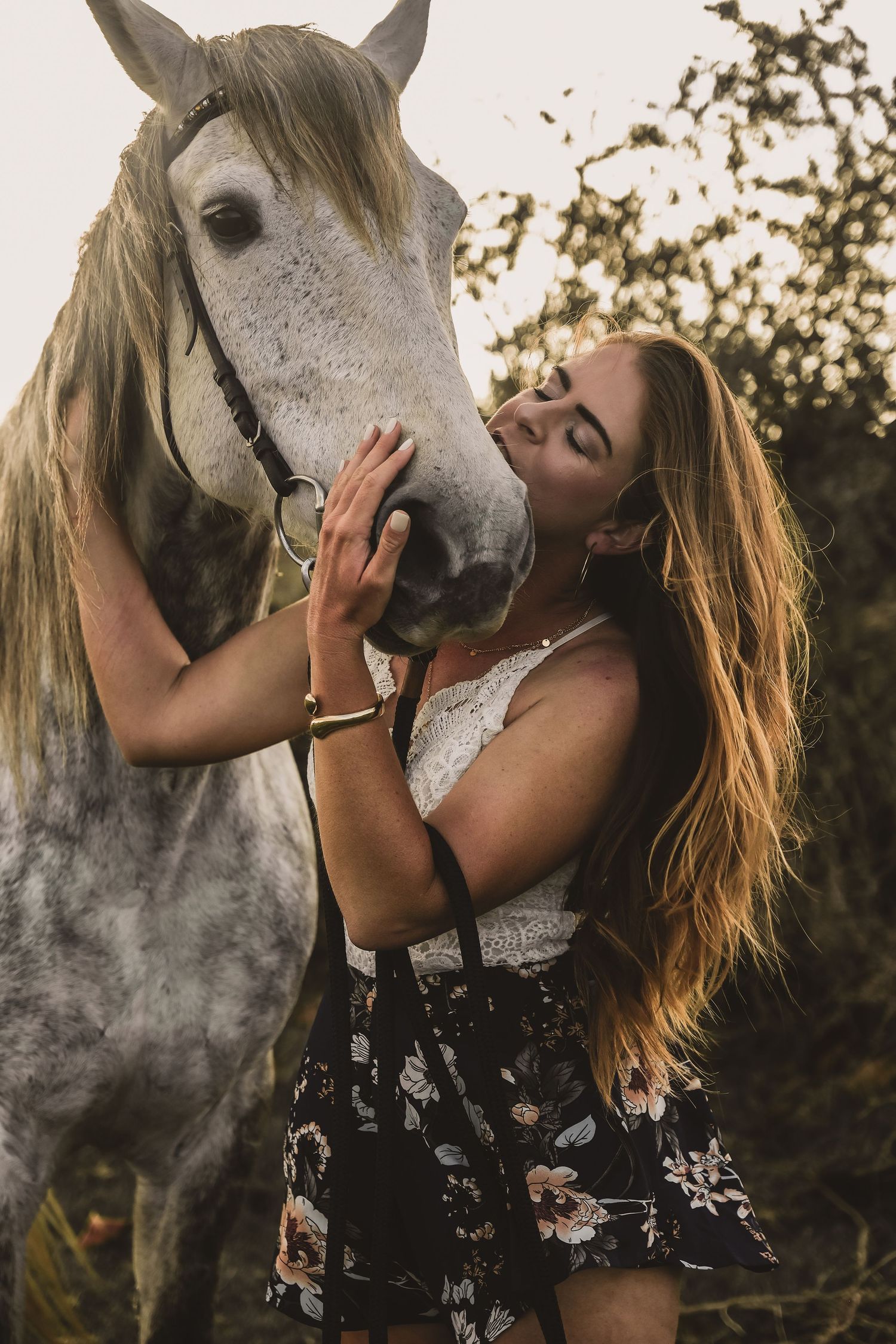 A dappled grey horse receives affection from someone in a flowing dress during a golden sunset.
