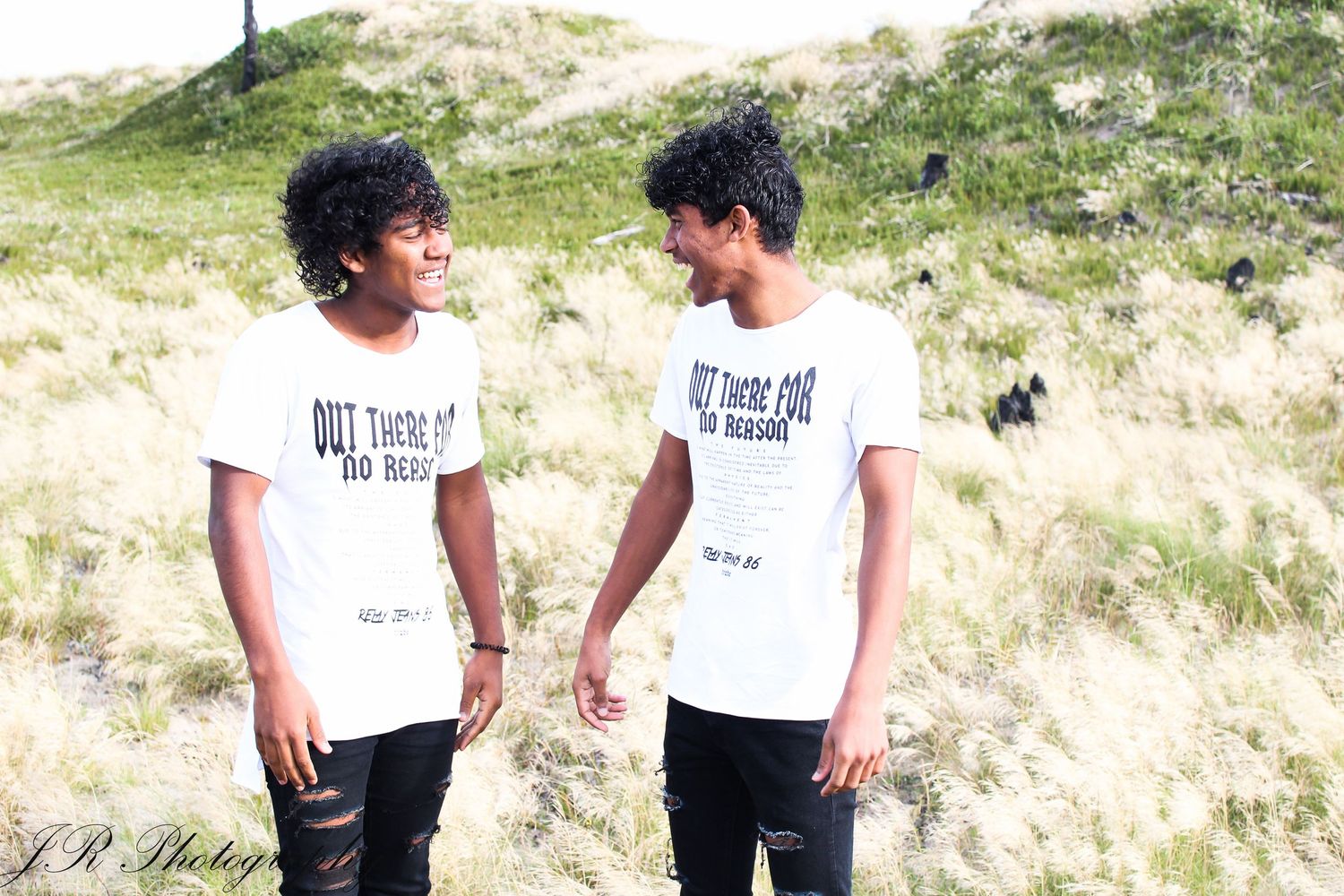 Two people in matching white t-shirts stand together in a sunny field with tall grass.