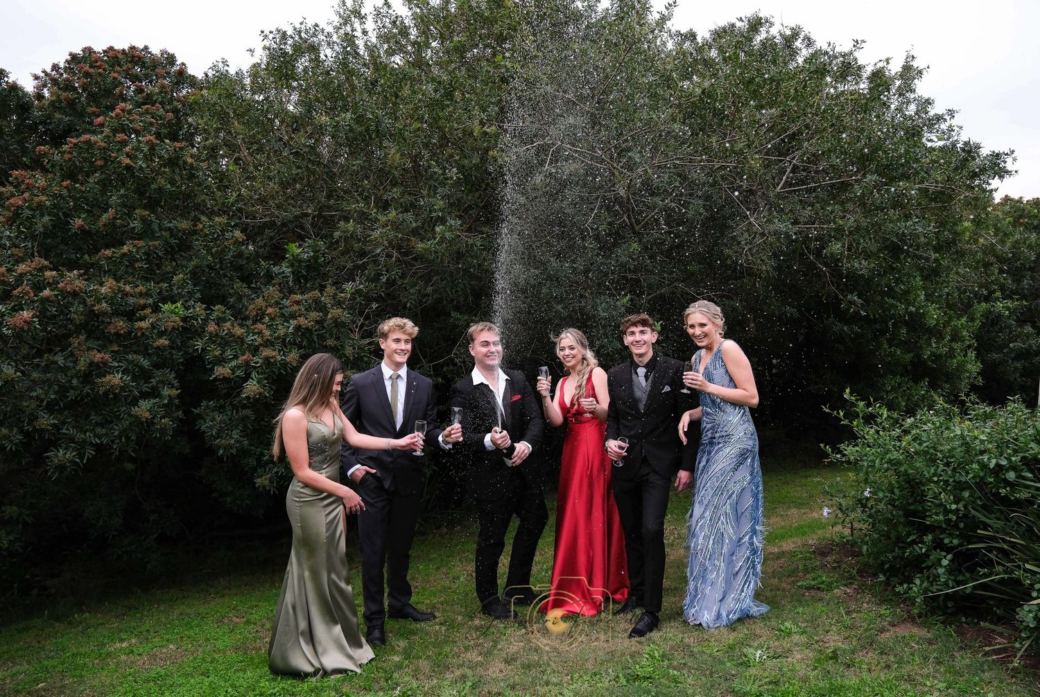 A group in formal attire celebrating with champagne spraying in a garden setting.