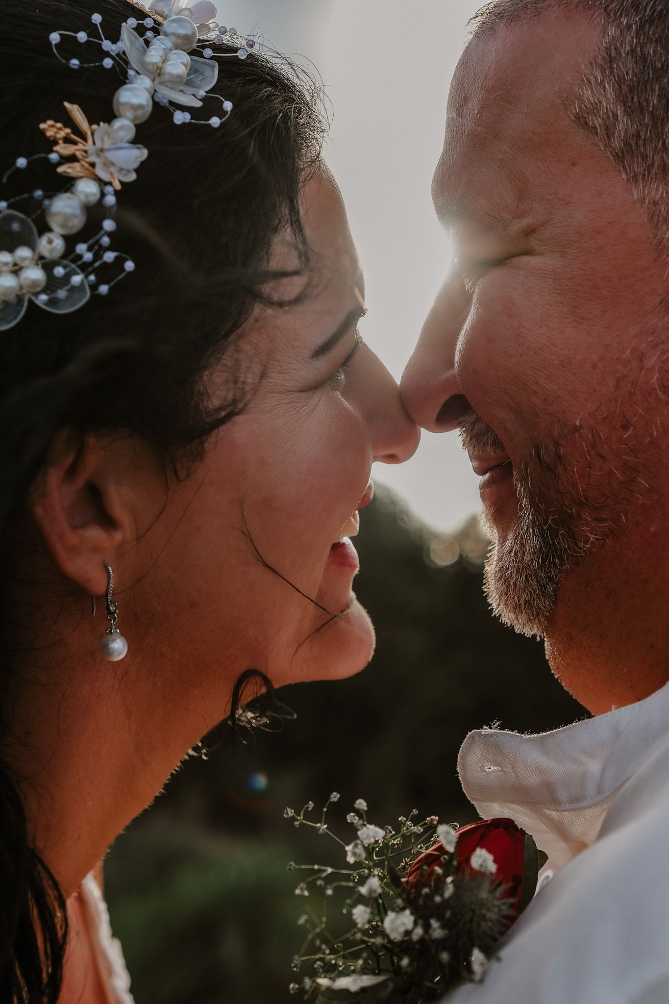 Close-up romantic wedding portrait with delicate floral crown and soft sunlight glowing behind the happy couple.