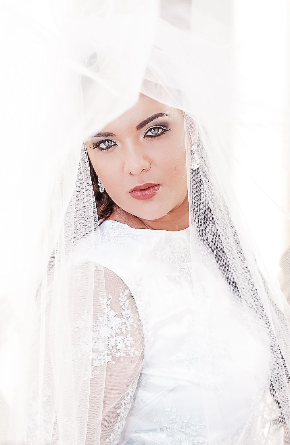 A bride wearing a white lace dress and delicate veil in a dreamy, high-key portrait.