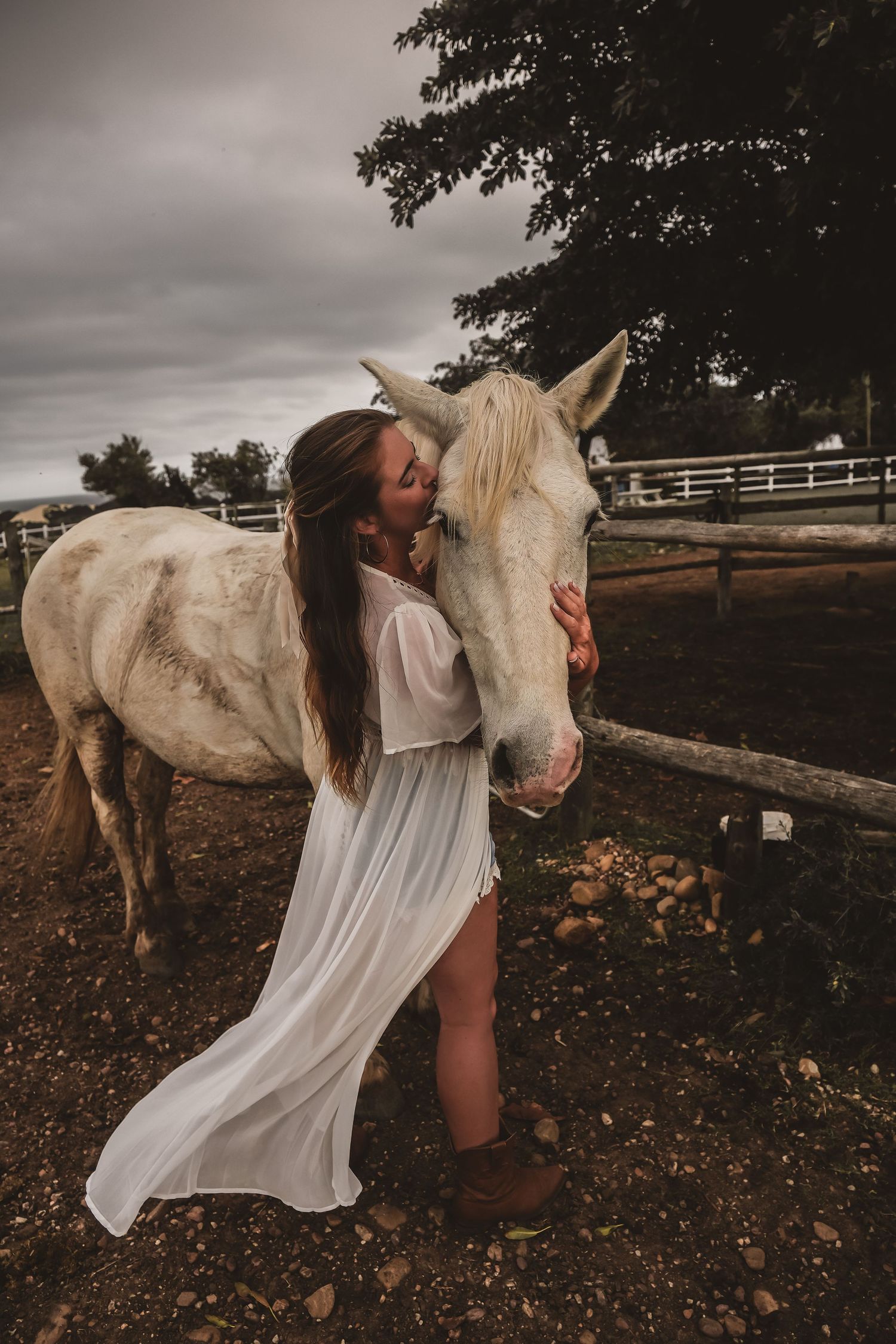 Woman in flowing white dress embraces white horse in rustic countryside setting.