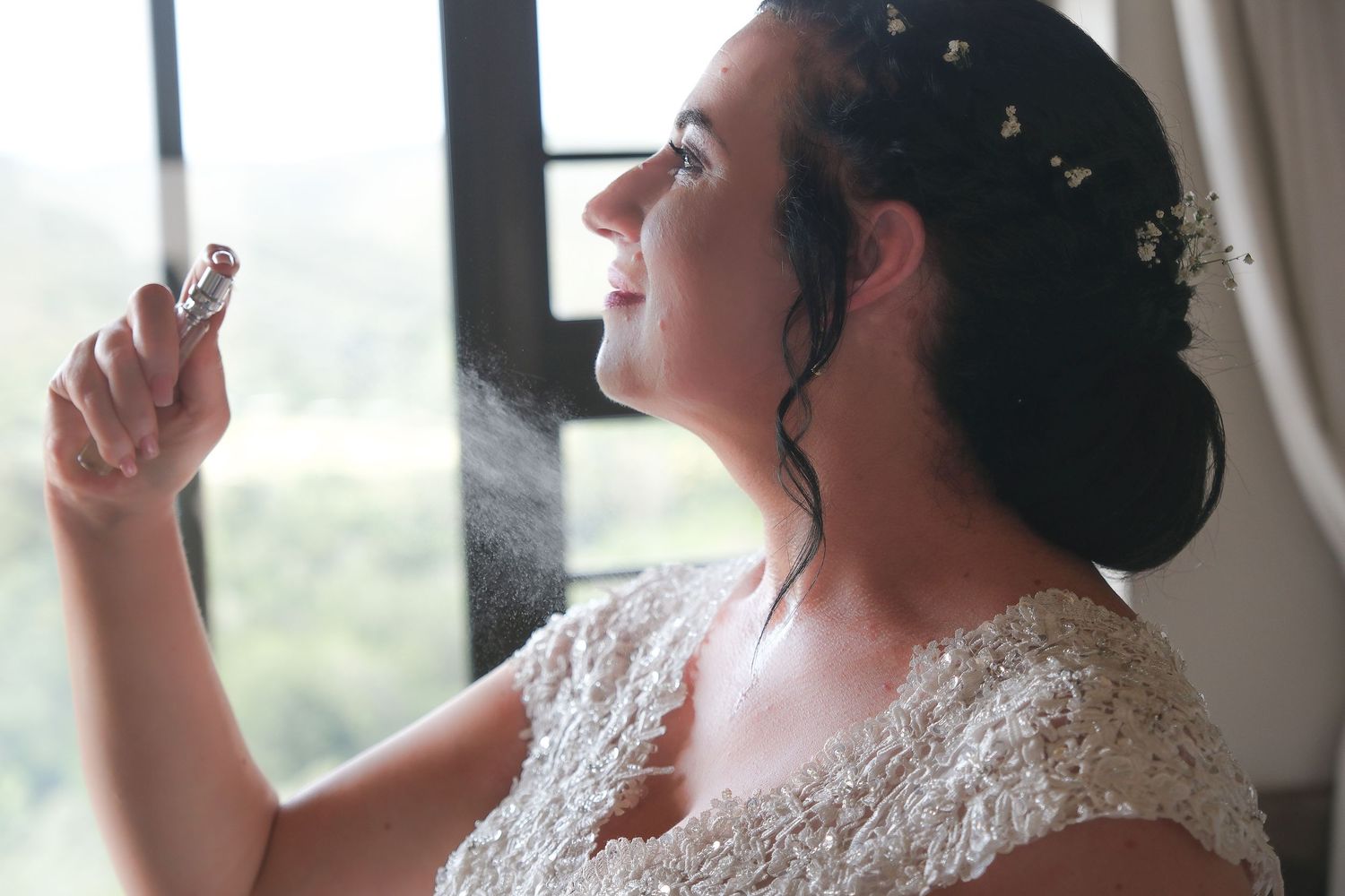 Bride in lace dress looking up thoughtfully by window with soft natural light.