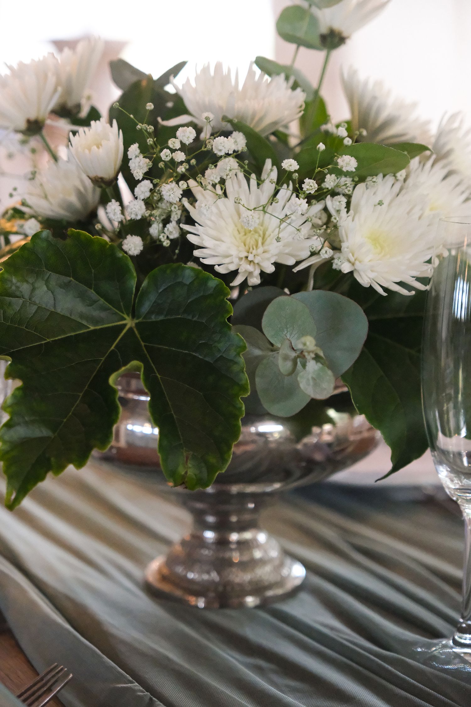 White chrysanthemums and baby's breath in a silver vase with eucalyptus leaves on a gray tablecloth.