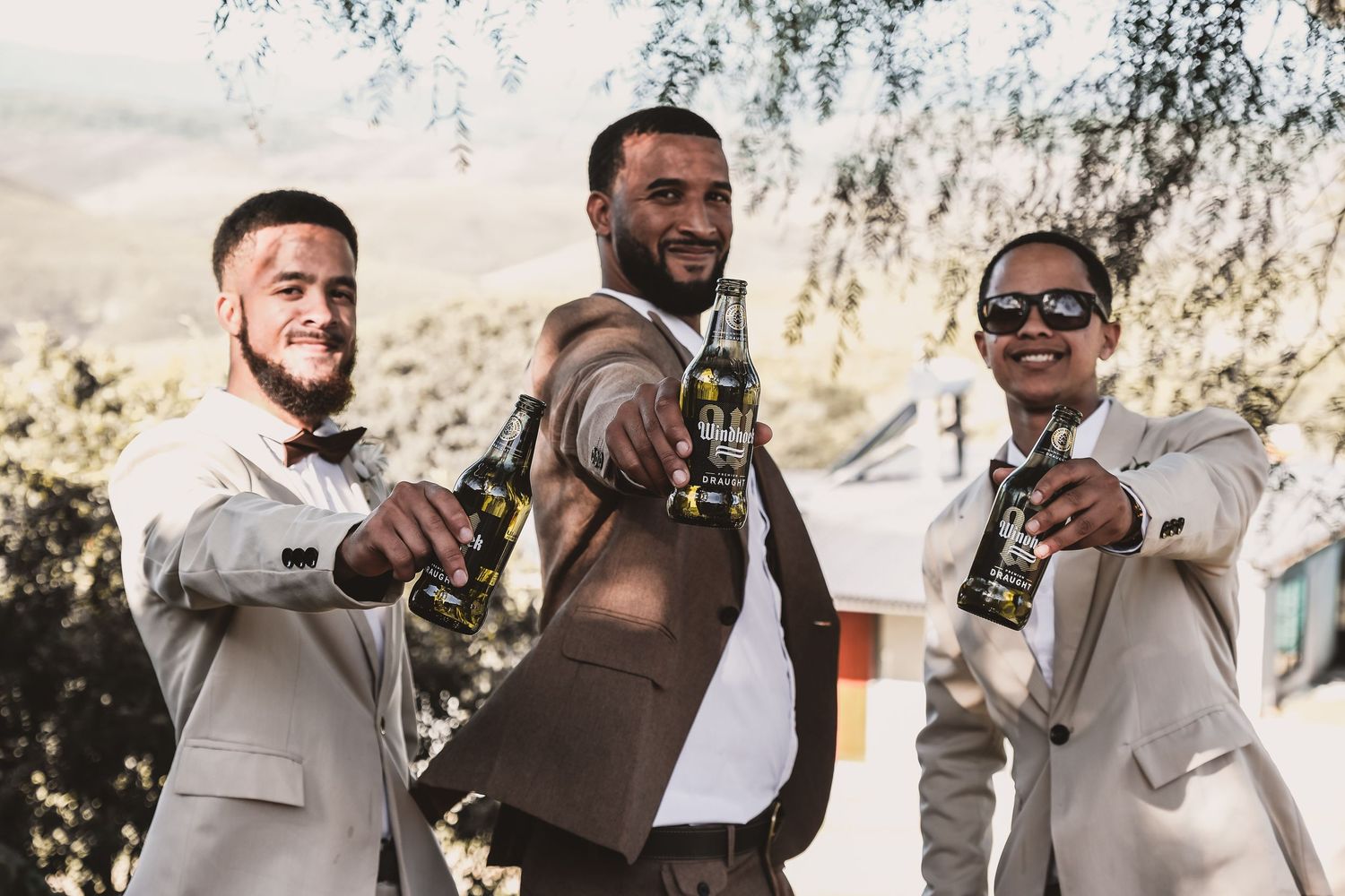 Three groomsmen in light colored suits toast with bottles during an outdoor celebration.