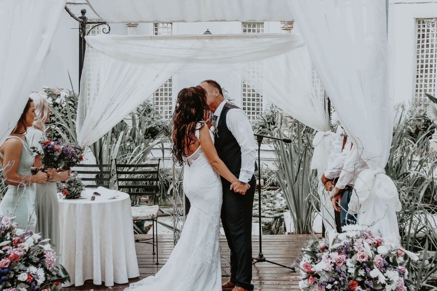 Elegant outdoor wedding ceremony under white draped arch with pink and purple floral arrangements.