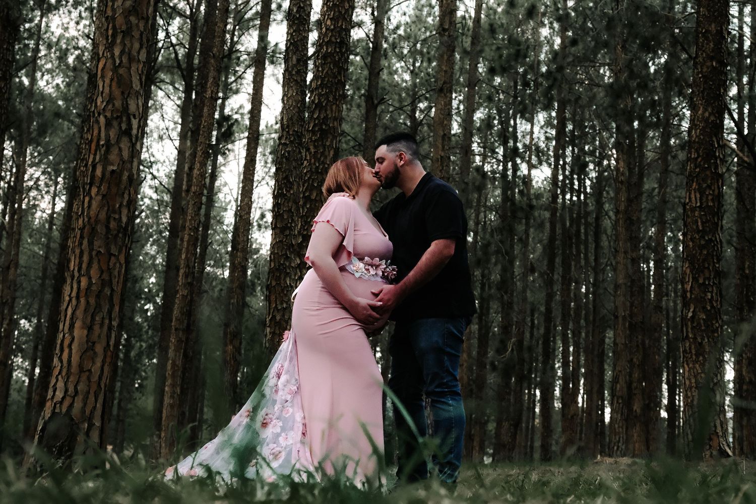 A couple stands intimately together in a moody forest setting with tall pine trees surrounding them.