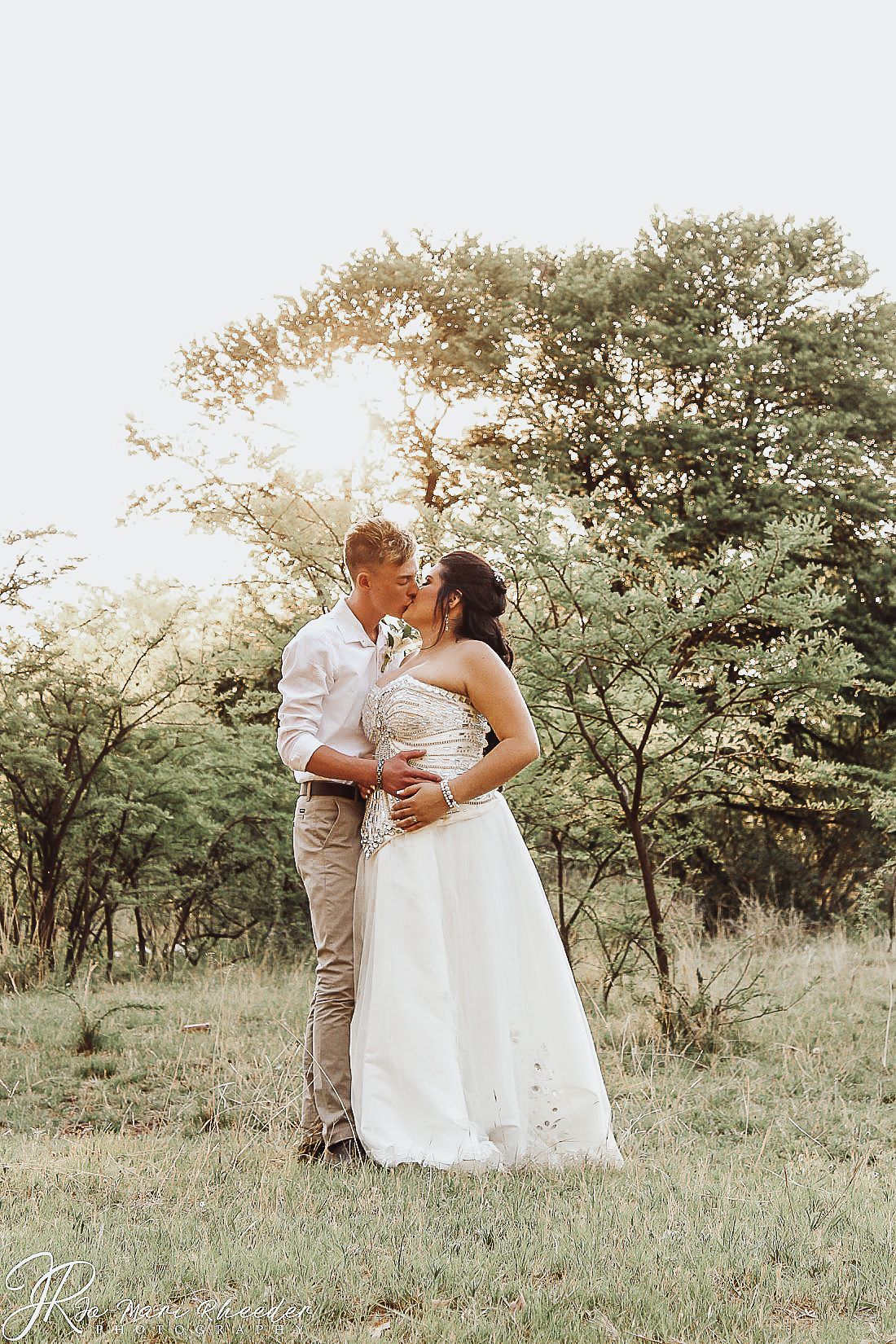 Romantic outdoor wedding portrait with sunlight glowing through trees in rustic field setting.