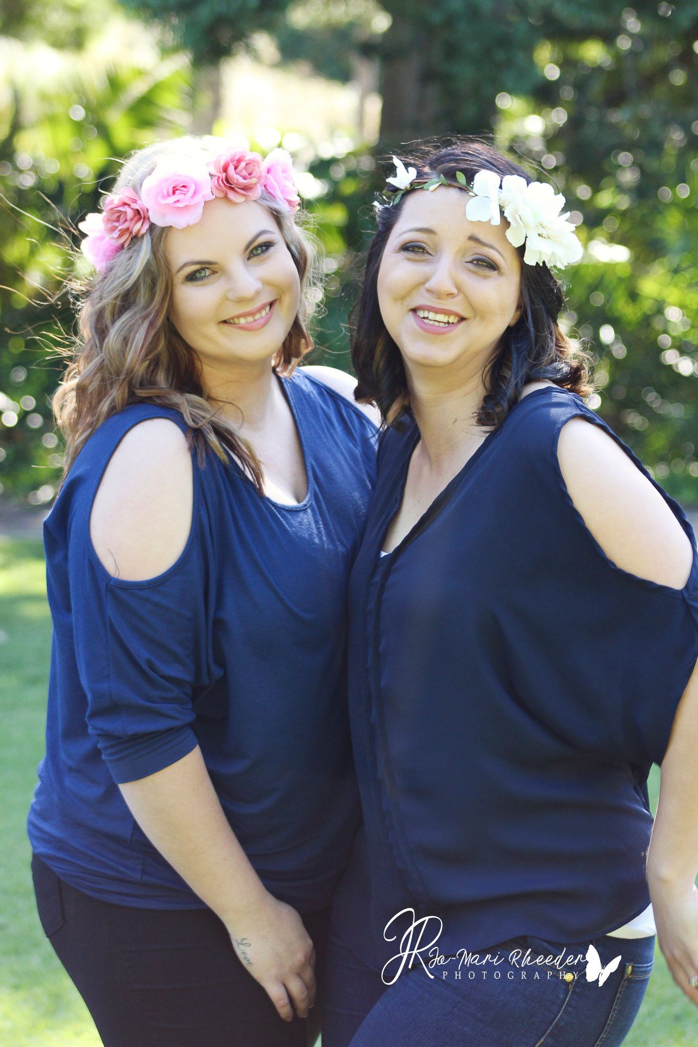 Two women in navy tops wearing flower crowns pose together outdoors.