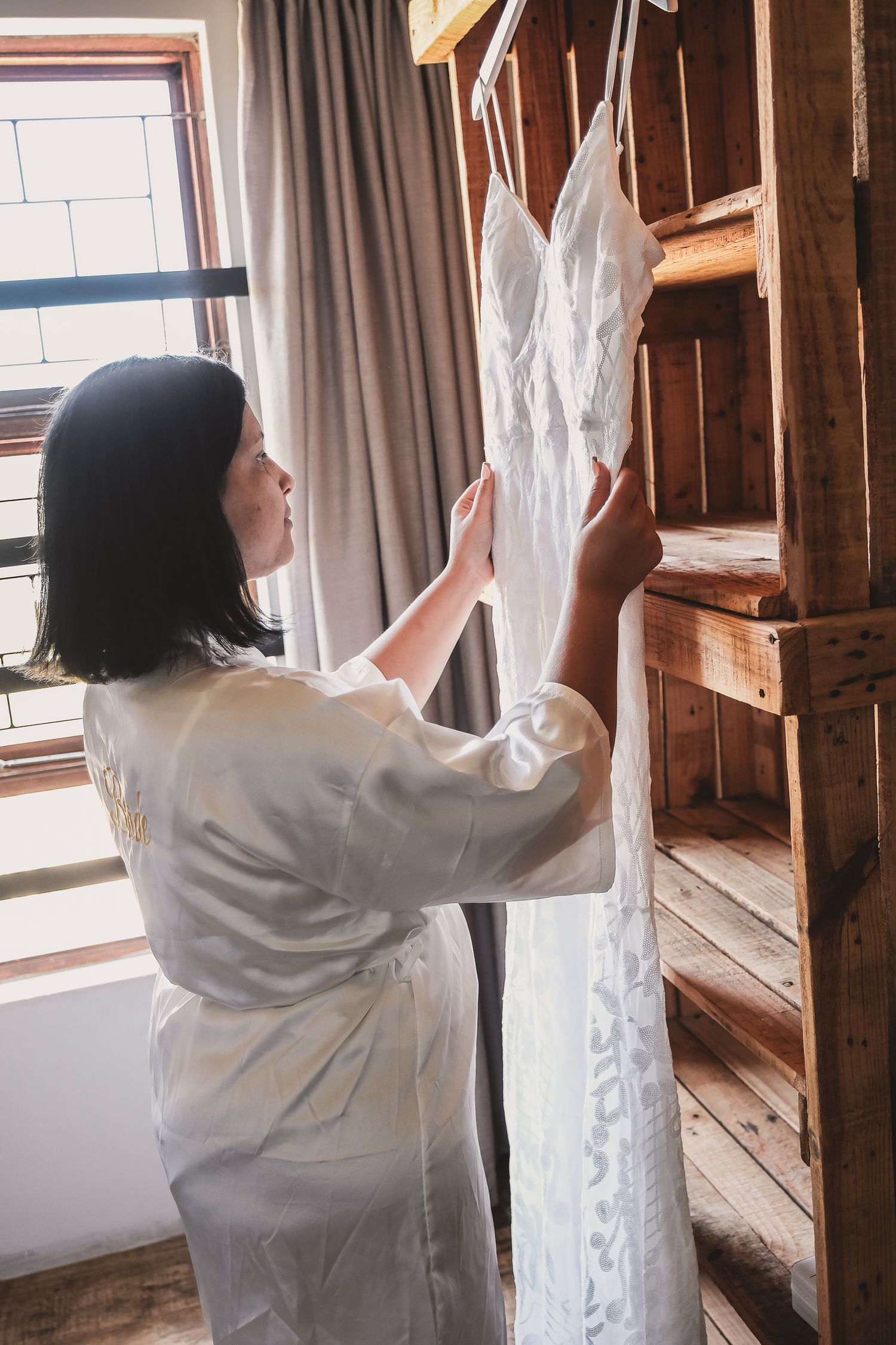 Person in white robe handling white wedding dress hanging on wooden wardrobe.