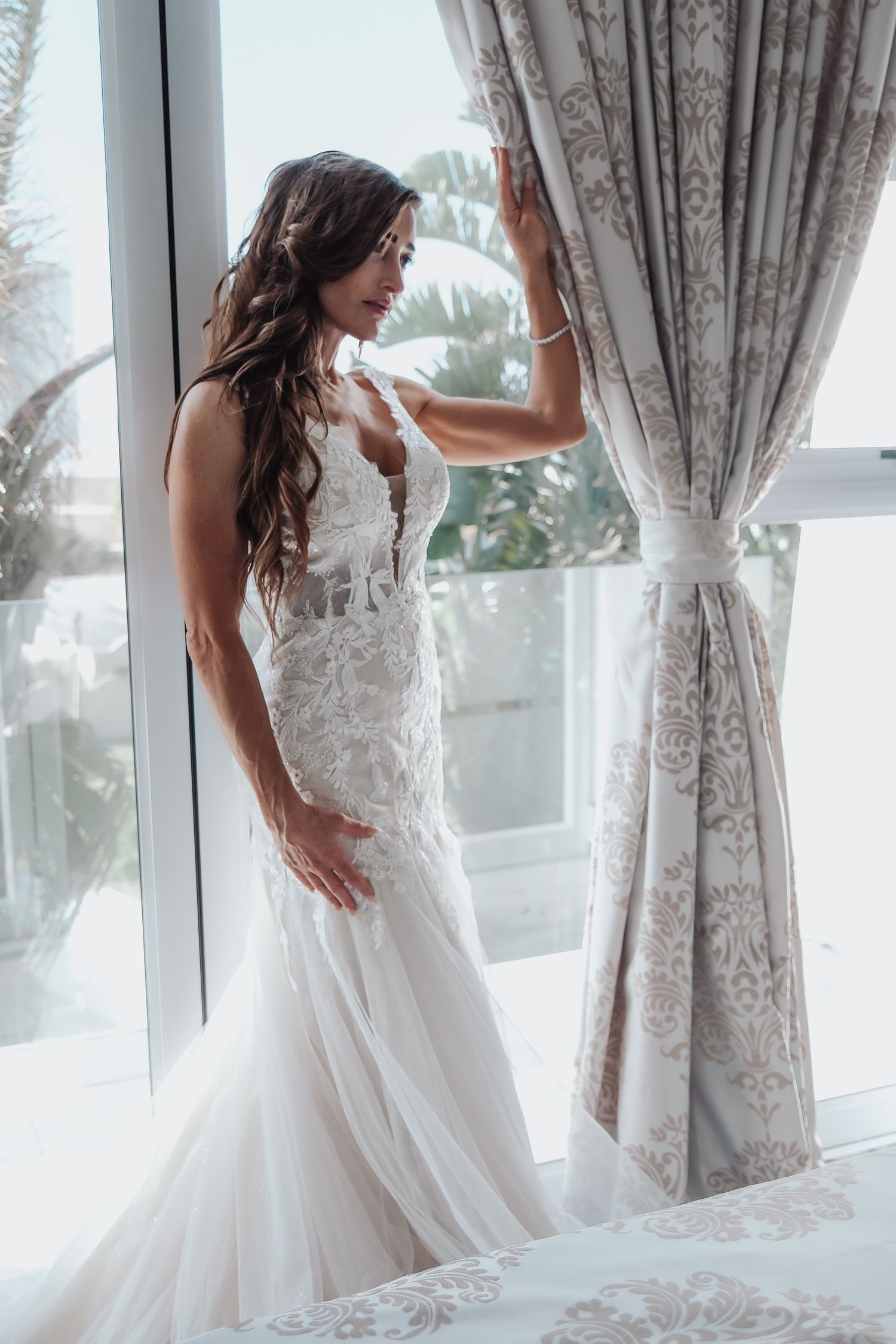 Bride in lace mermaid wedding dress poses elegantly by window with flowing curtains.