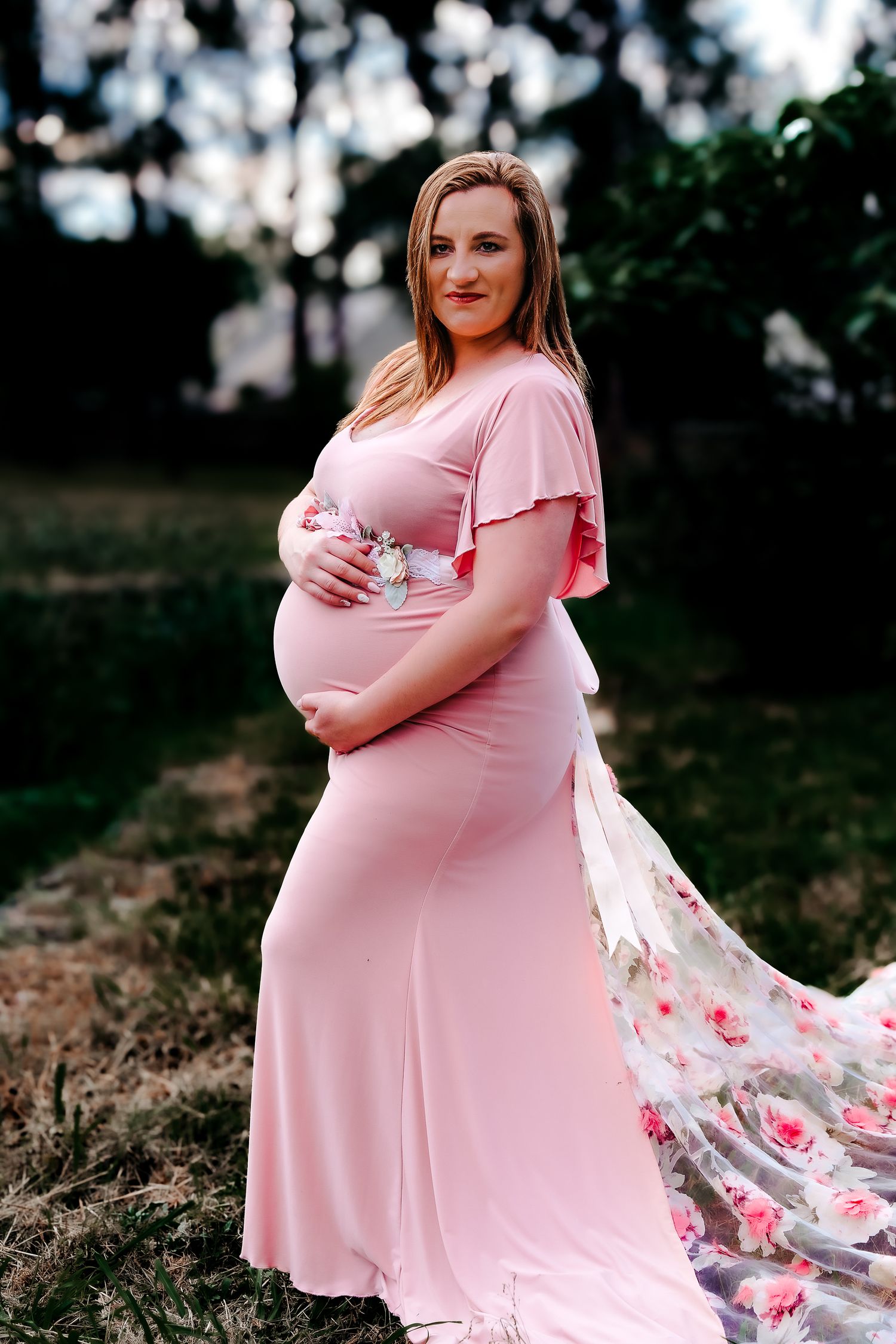 A pregnant woman in a pink maxi dress poses outdoors with a floral shawl flowing in the breeze at sunset.