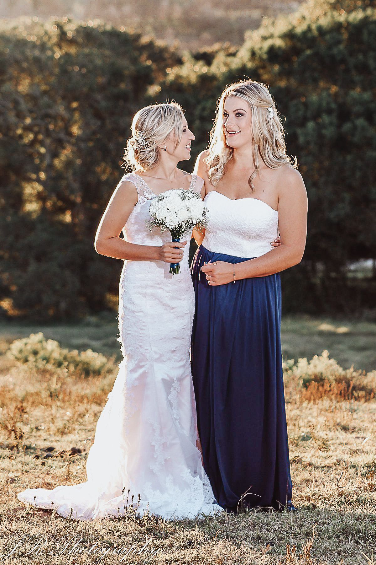 Wedding party members in elegant white lace gown and long navy blue dress pose together outdoors at sunset.