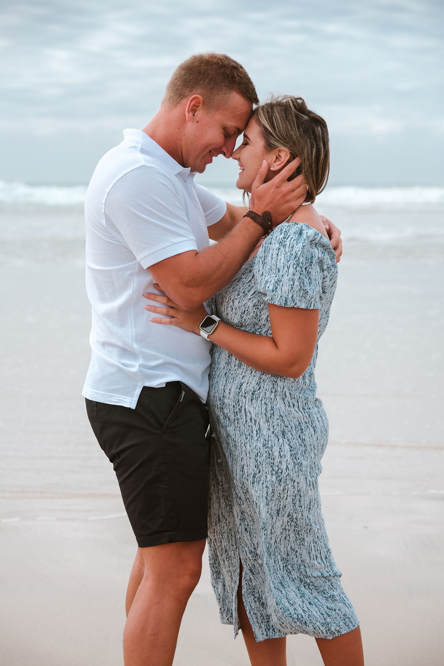 Two people share a romantic moment embracing on a beach at sunset in casual summer attire.