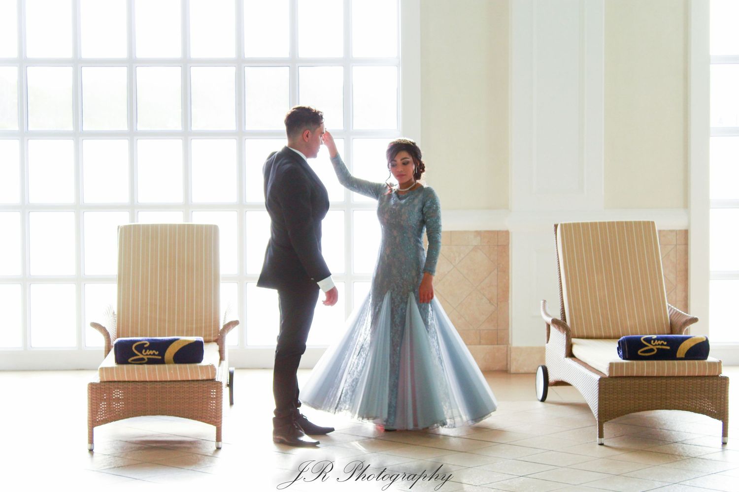 Two people share an intimate moment in formal attire against a bright window with lounge chairs.