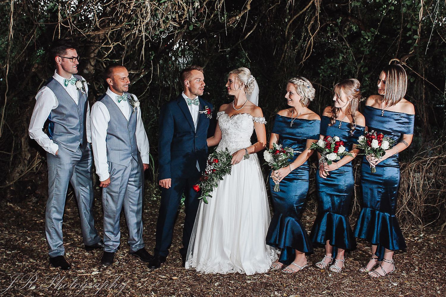 Wedding party poses together with bridesmaids in blue dresses against natural backdrop.