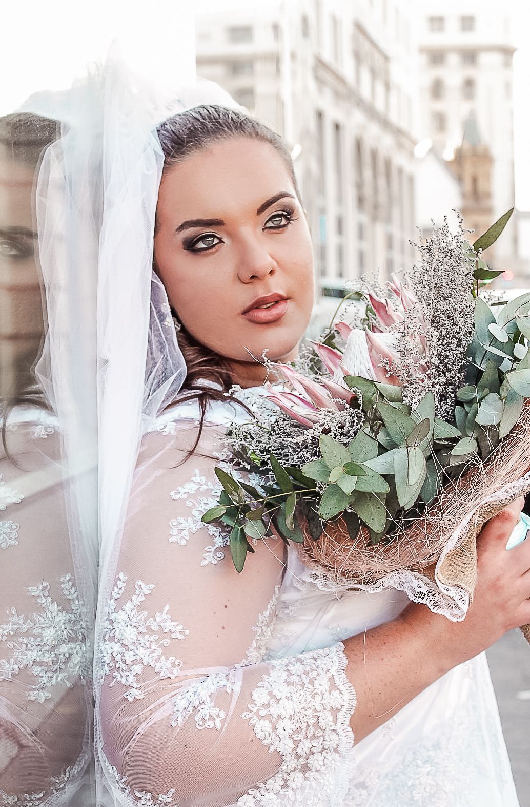 A bride in an elegant lace gown holding a eucalyptus bouquet against an urban backdrop.