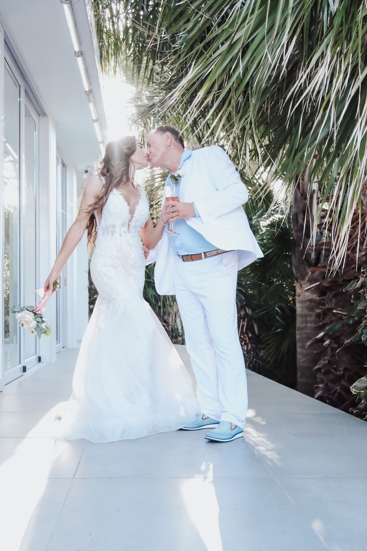 Sunlit wedding couple kissing under palm trees in all-white attire.