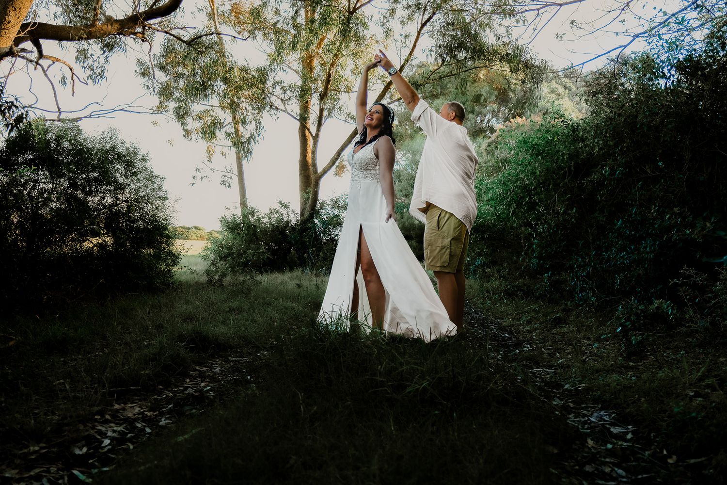 Two people in white outfits dancing together in a dreamy outdoor woodland setting during golden hour.