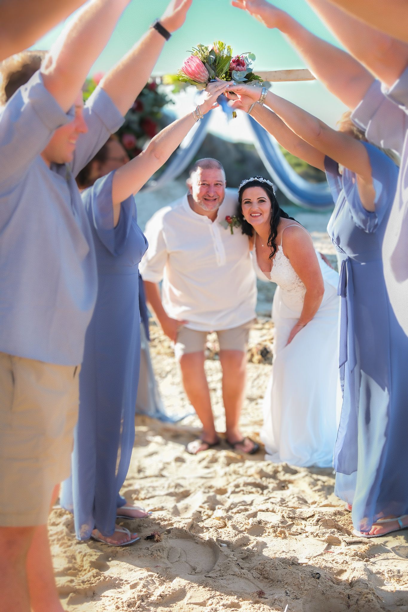A couple walk through an arch formed by bridesmaids holding bouquets on a sunny beach during their wedding ceremony.