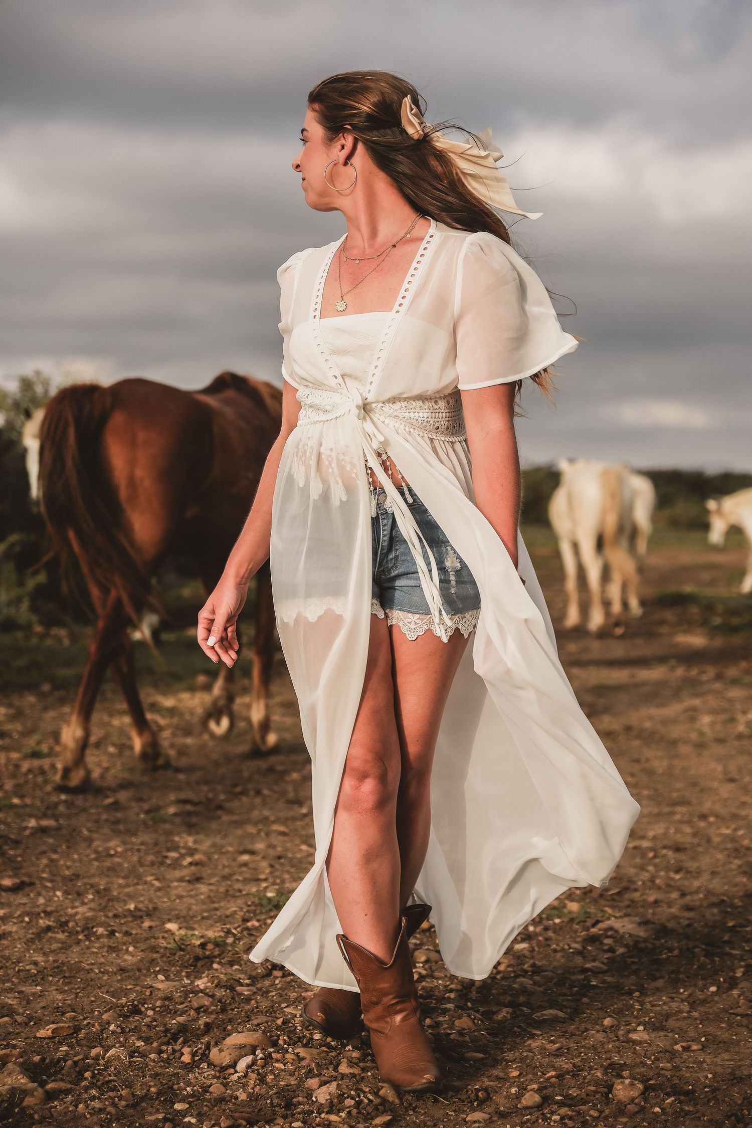 Someone in a flowing white dress walks through a field with horses grazing in the background.