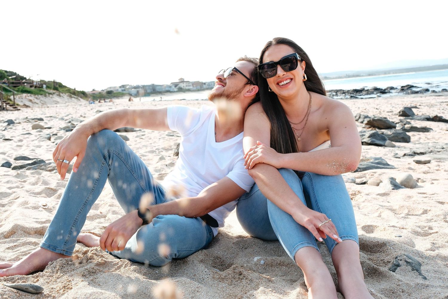 Two people relaxing on the beach in casual denim attire and sunglasses.
