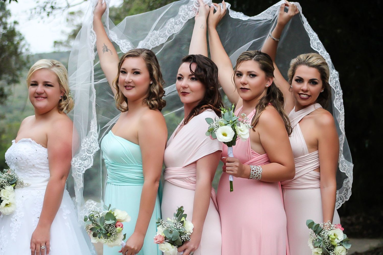 Bridal party in pastel dresses poses together while holding a flowing veil.