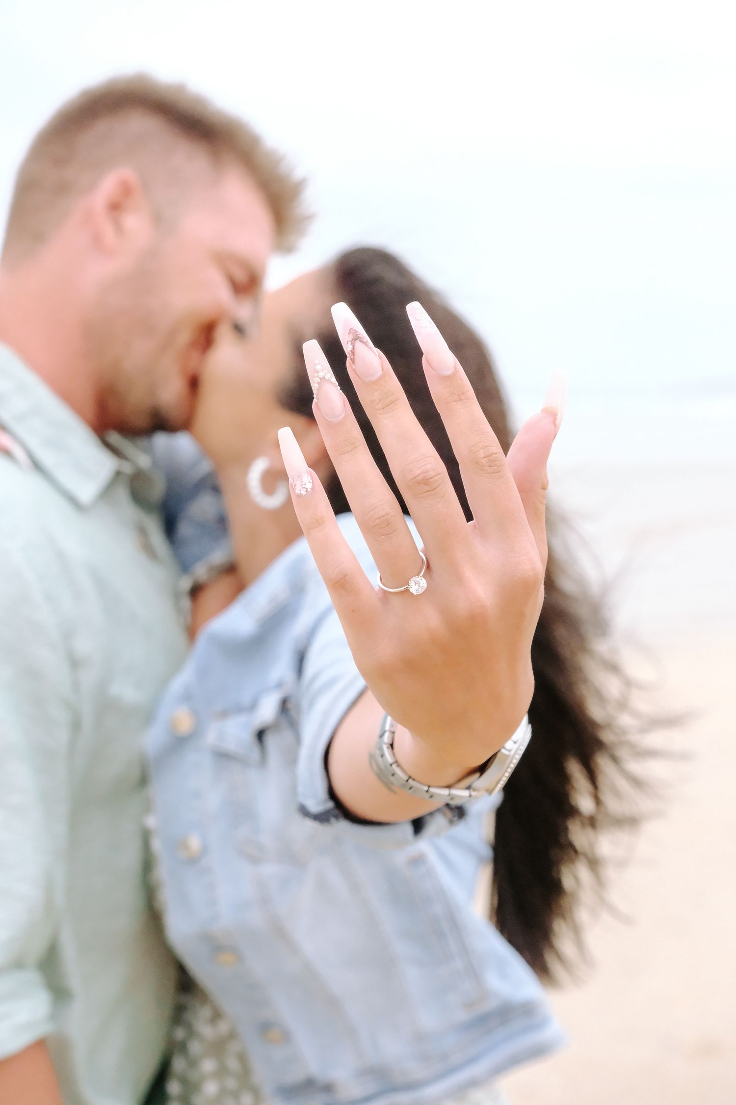 An engagement ring sparkles as a couple embraces in soft focus background.