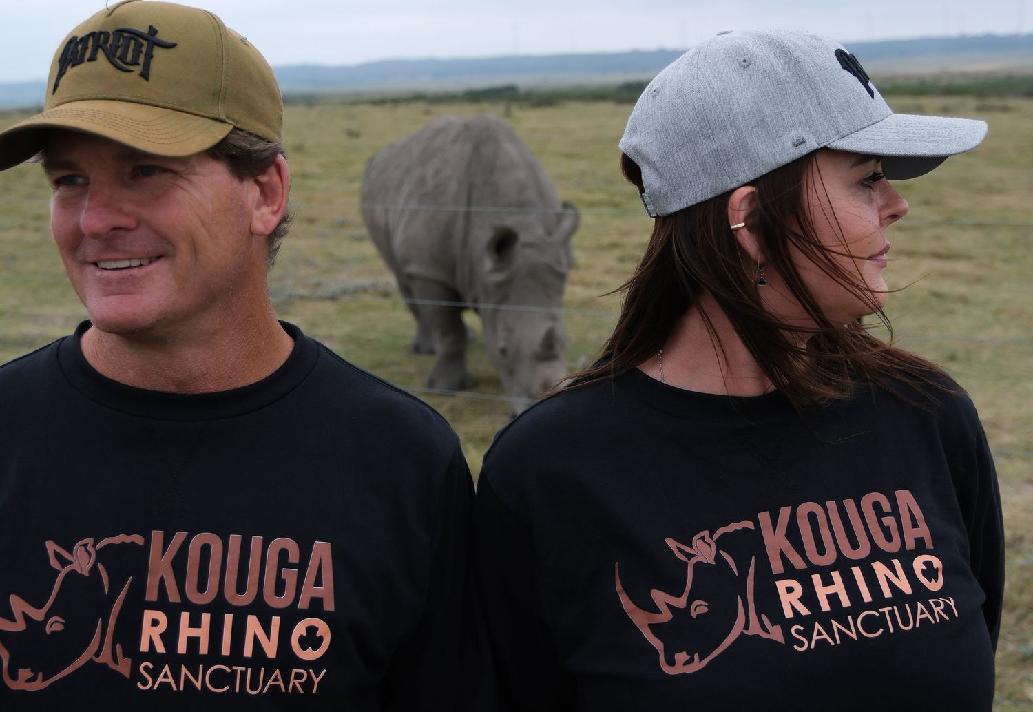 Two people wearing Kouga Rhino Sanctuary shirts pose with a rhino in the background.