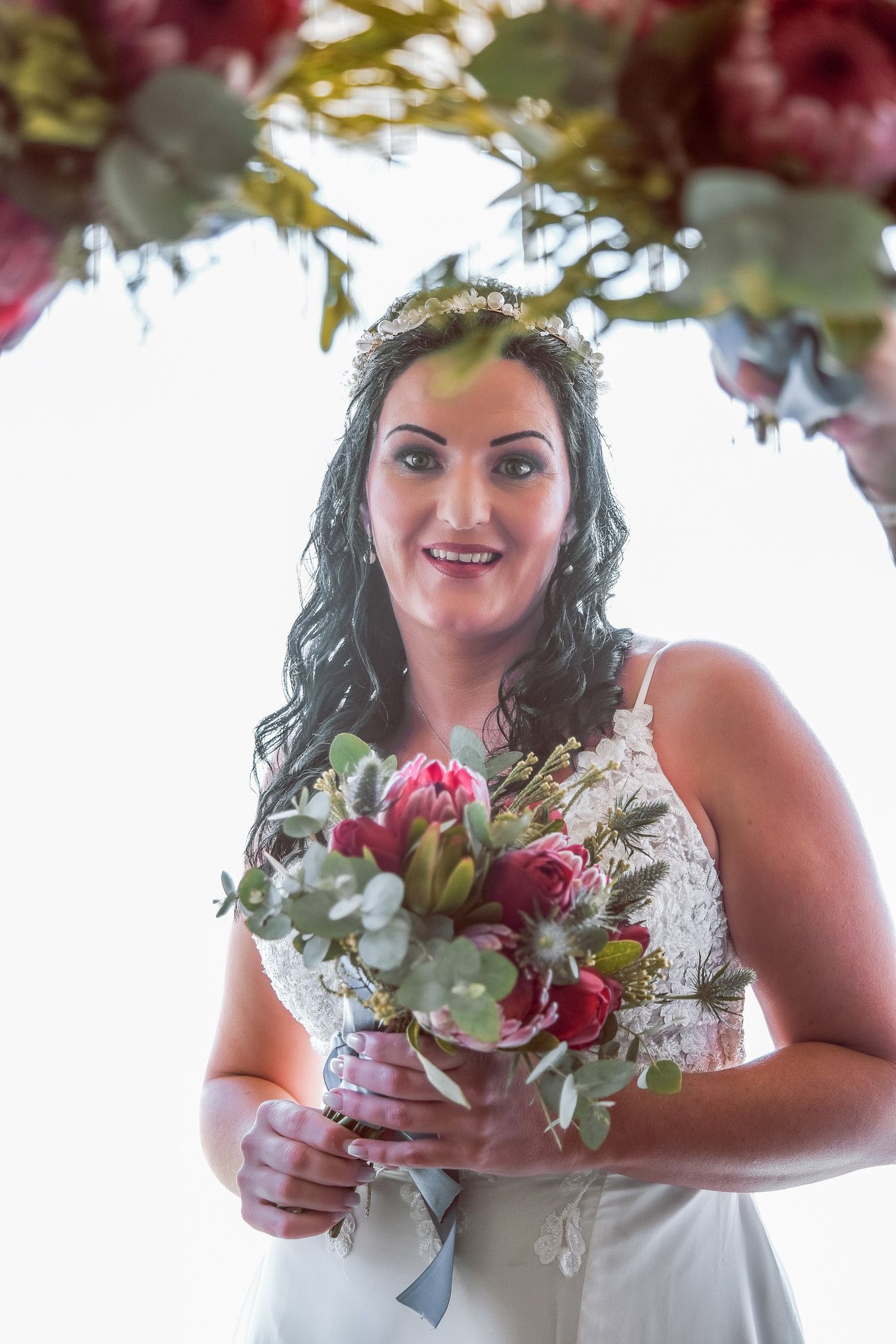 A bride in a white dress holding a vibrant bouquet of pink and red flowers stands beneath a floral arch.