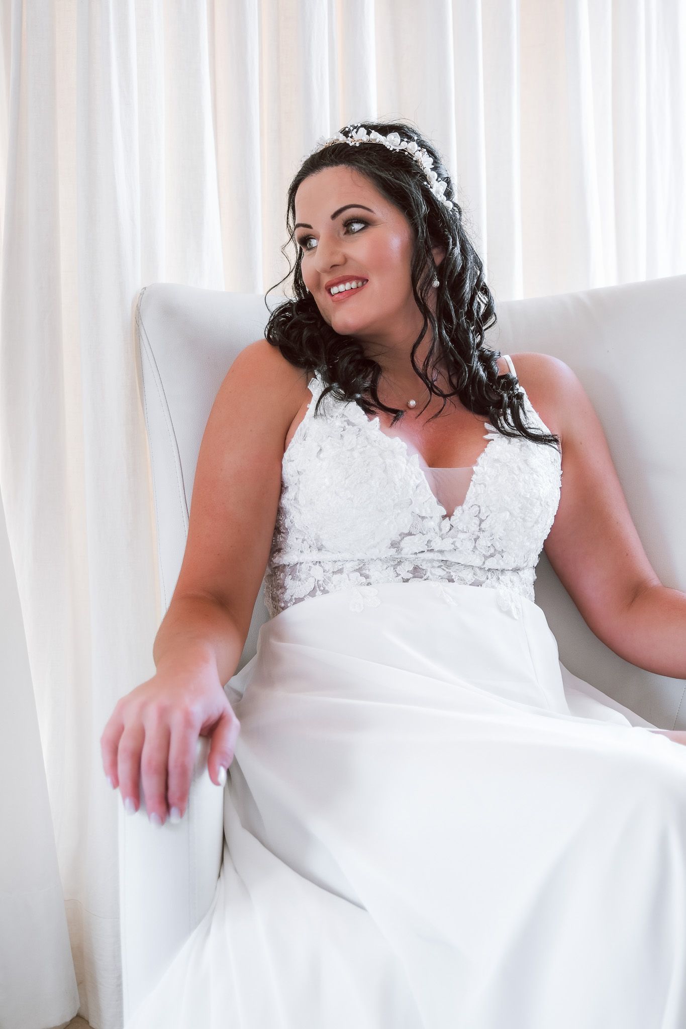 A bride wearing a white sleeveless wedding gown with lace detail sits elegantly in a white armchair.