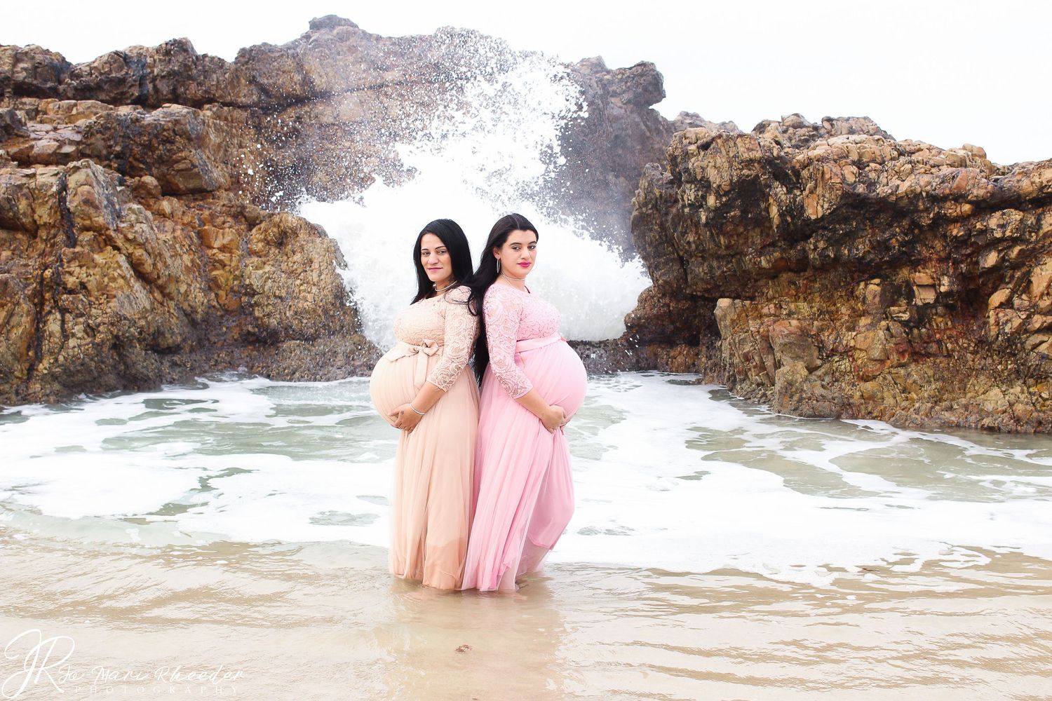 Two expectant mothers in flowy gowns stand together on a beach with dramatic rocky coastline backdrop.