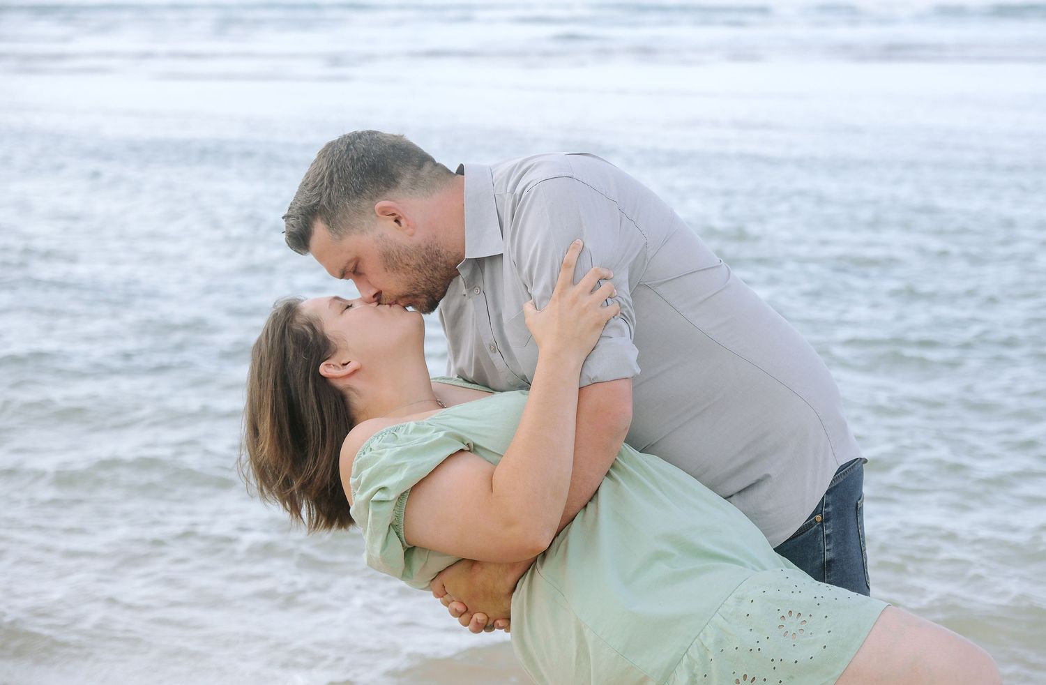 A romantic couple embraces on a beach with waves in the background during a photoshoot.