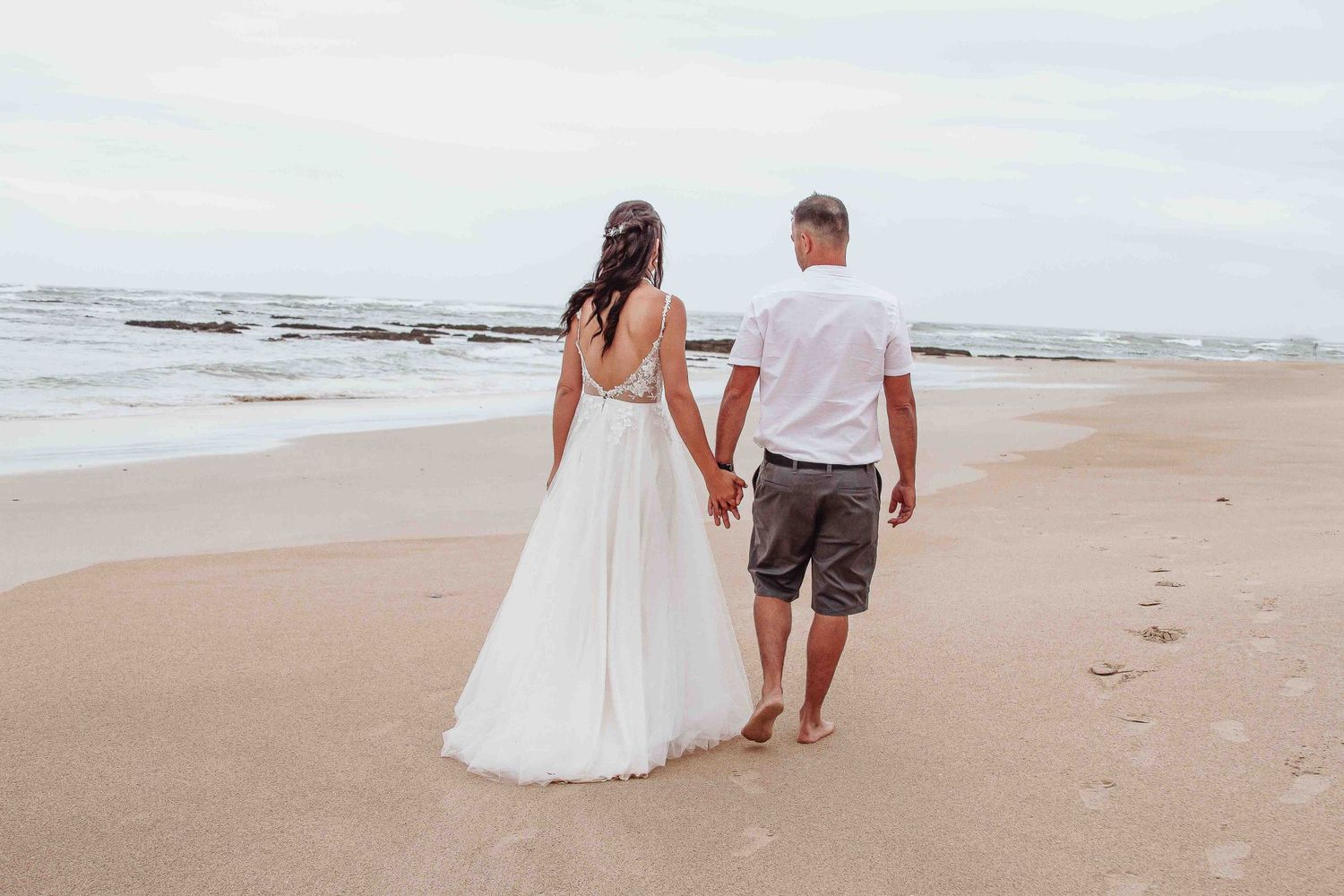 A couple walks hand in hand along the shoreline in wedding attire with footprints in the sand.